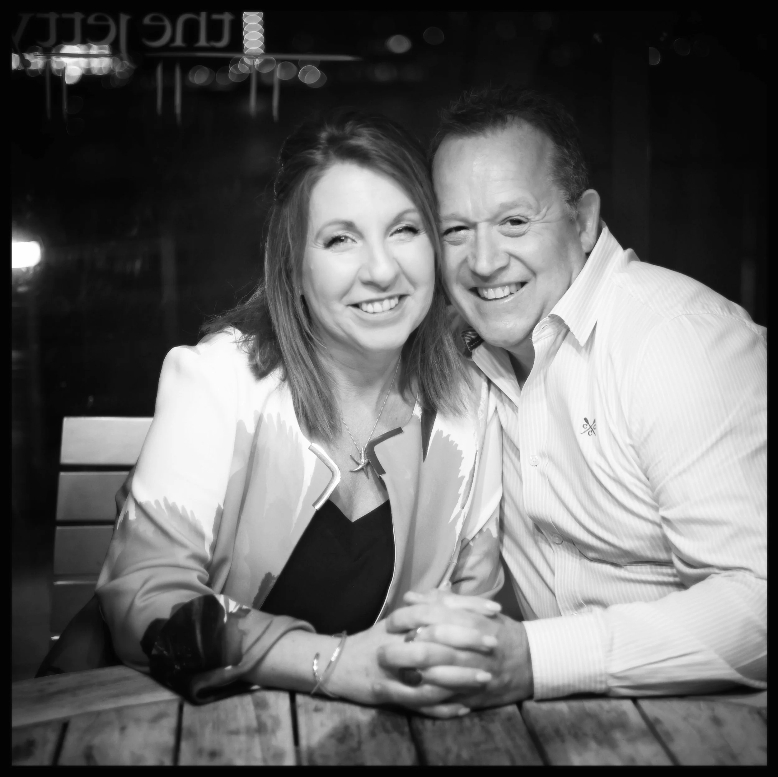 A happy couple smiling and leaning close together at a table, with the woman wearing a white dress and the man wearing a striped shirt, in a nighttime setting with bokeh lights in the background.