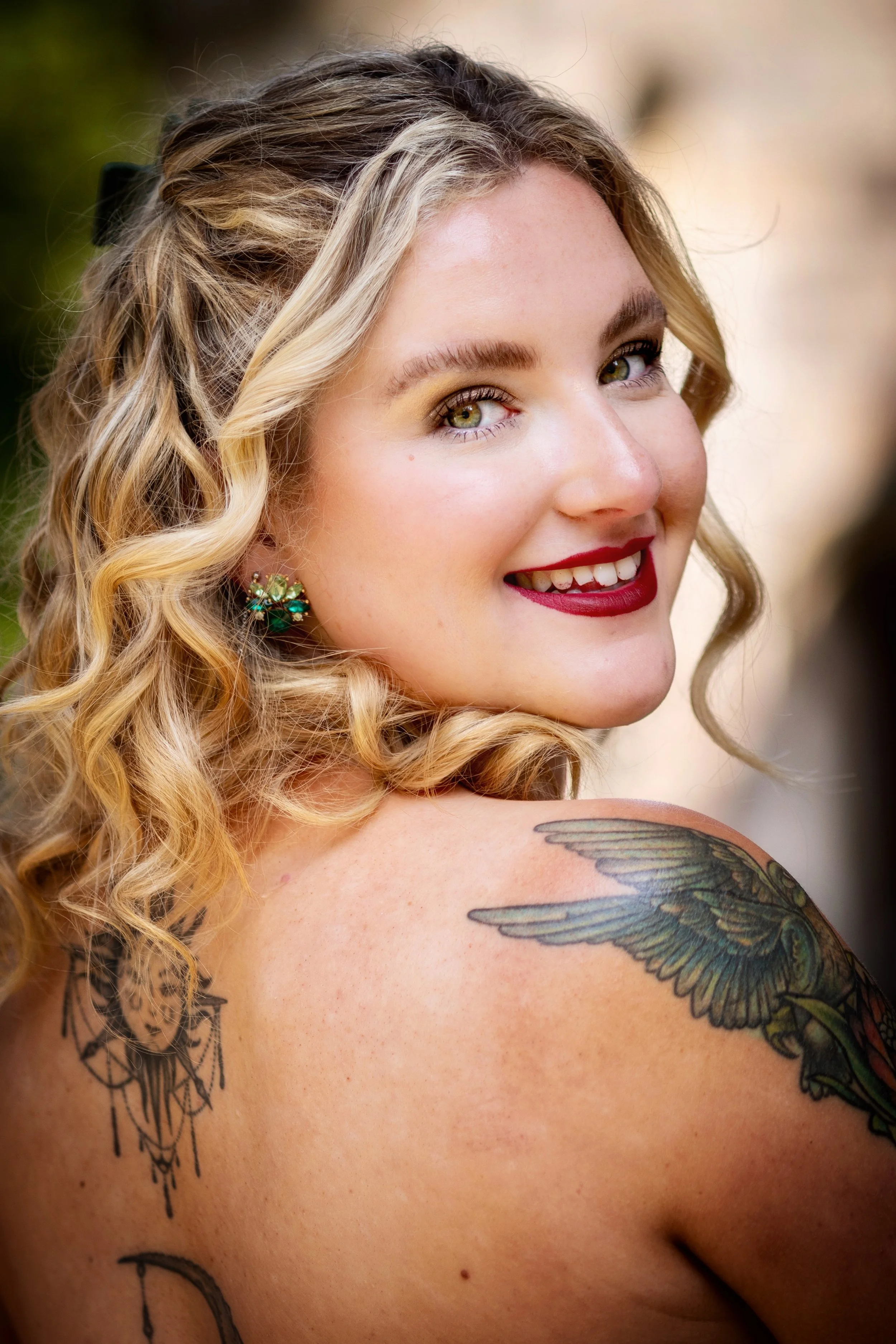 Close-up of a smiling woman with curly blonde hair, green eyes, red lipstick, gold earrings, and tattoos of a bird and lace design on her shoulder.