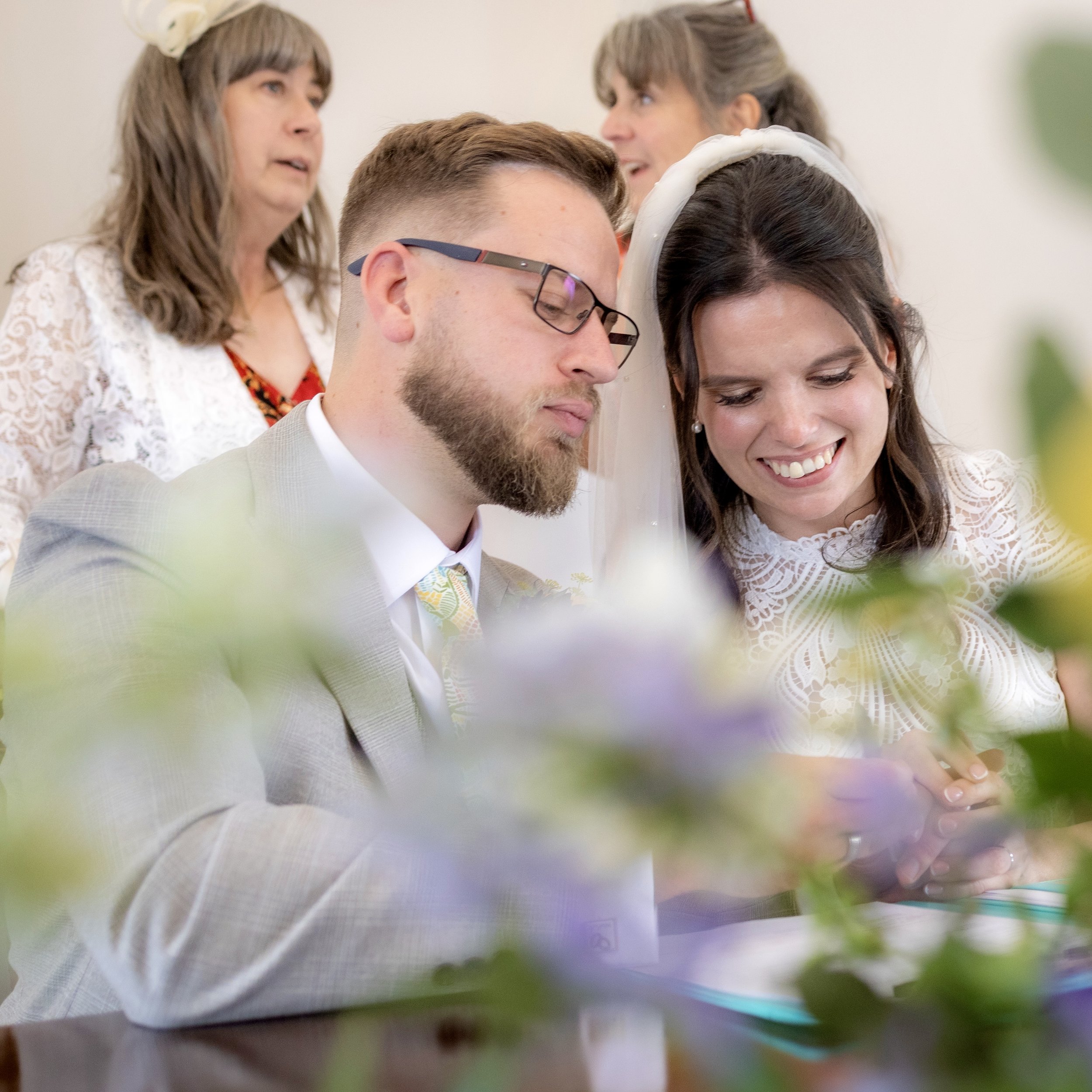 A couple at their wedding ceremony, signing documents, surrounded by family members, with green foliage in the foreground.