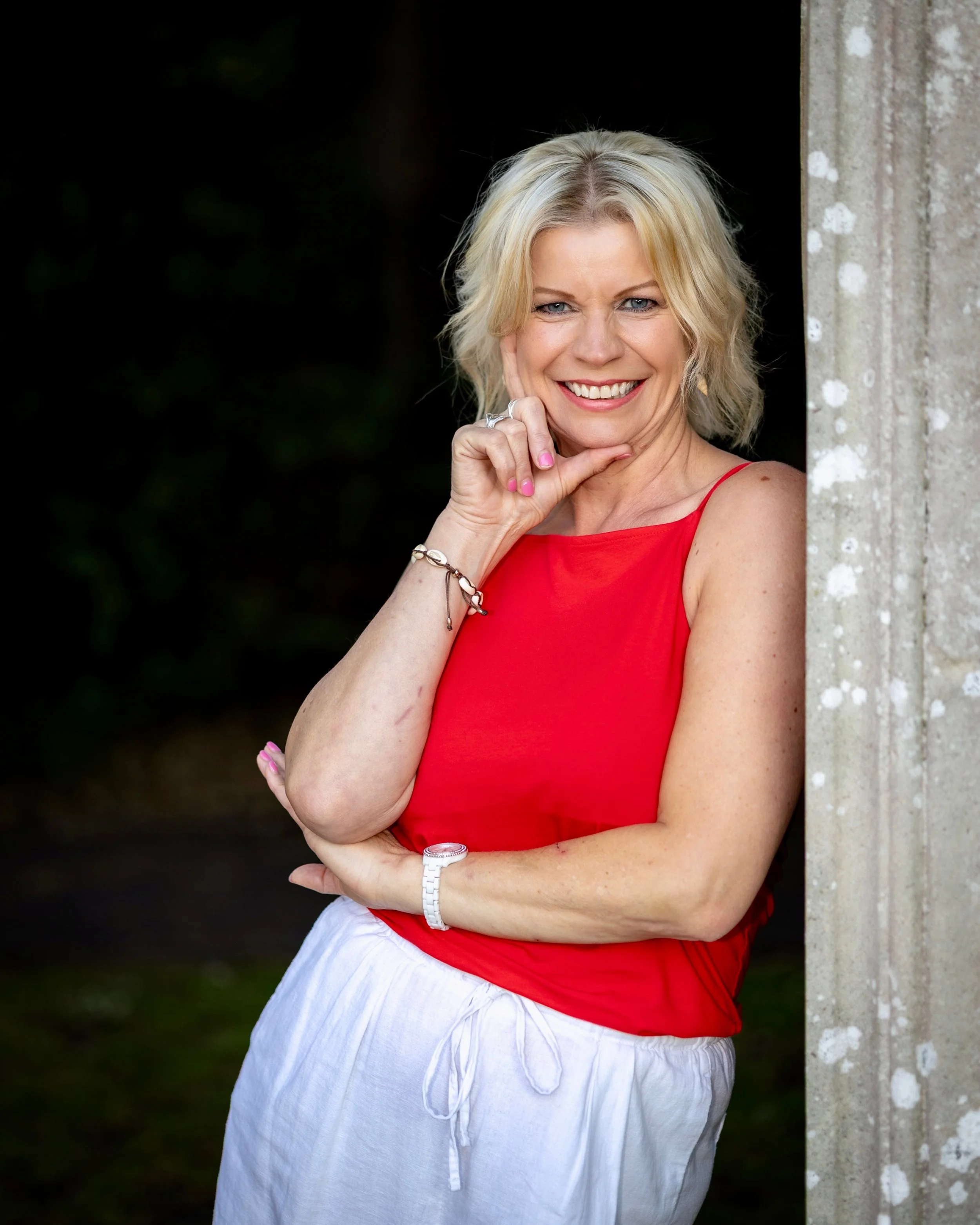 A woman with blonde hair wearing a red sleeveless top and white pants stands outdoors near a concrete pillar, smiling and posing with her hand on her chin in front of a dark, blurred background.