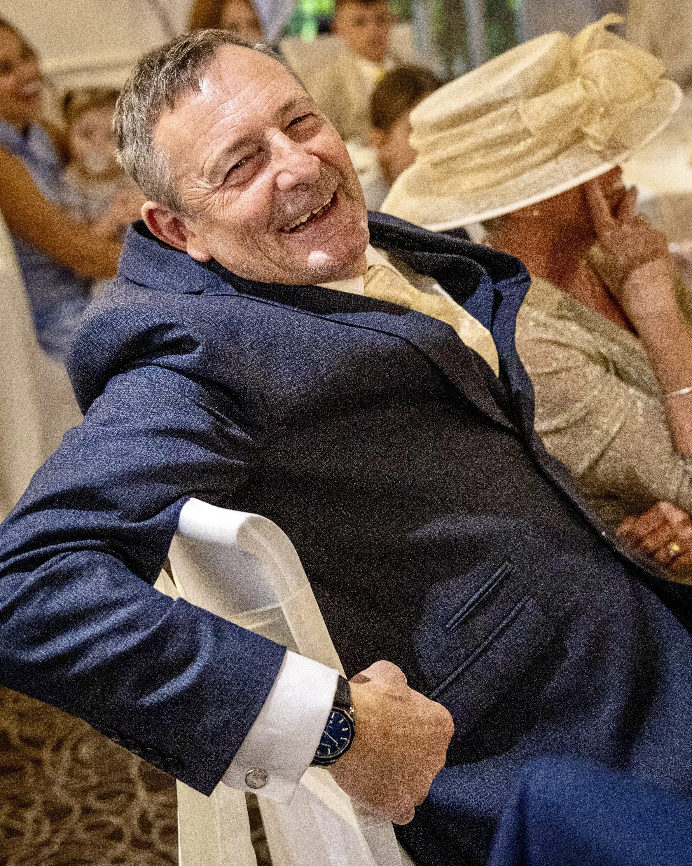 Close-up photo of a man smiling and wearing a dark suit with a white shirt, sitting at a table with people in the background, one woman with a large beige hat.