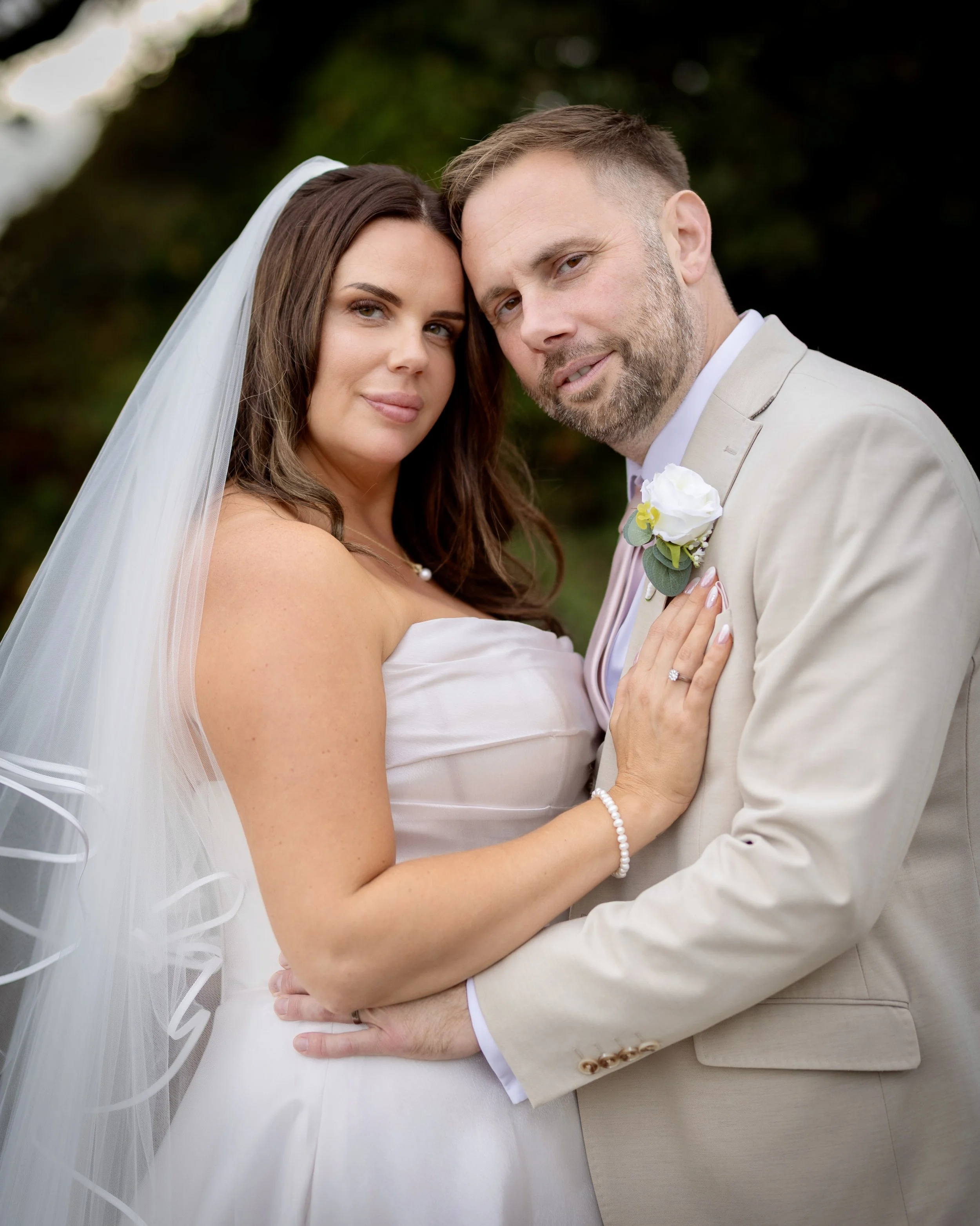 A bride and groom in wedding attire embrace outdoors in front of a dark background with greenery. The bride wears a white dress with thin straps, a pearl bracelet, and a veil. The groom wears a beige suit with a white shirt, a pink tie, and a boutonn