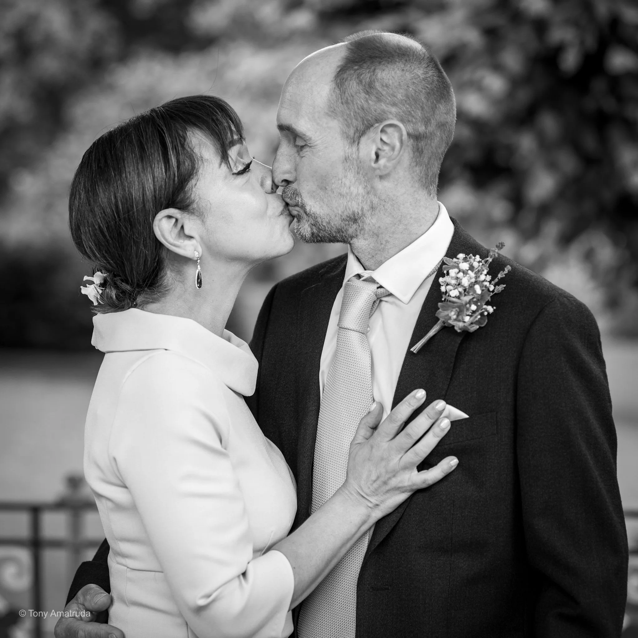 Black and white photograph of a couple sharing a kiss, with the woman wearing a white dress and dangling earrings, and the man in a suit with a boutonnière, outdoors with blurred foliage in the background.