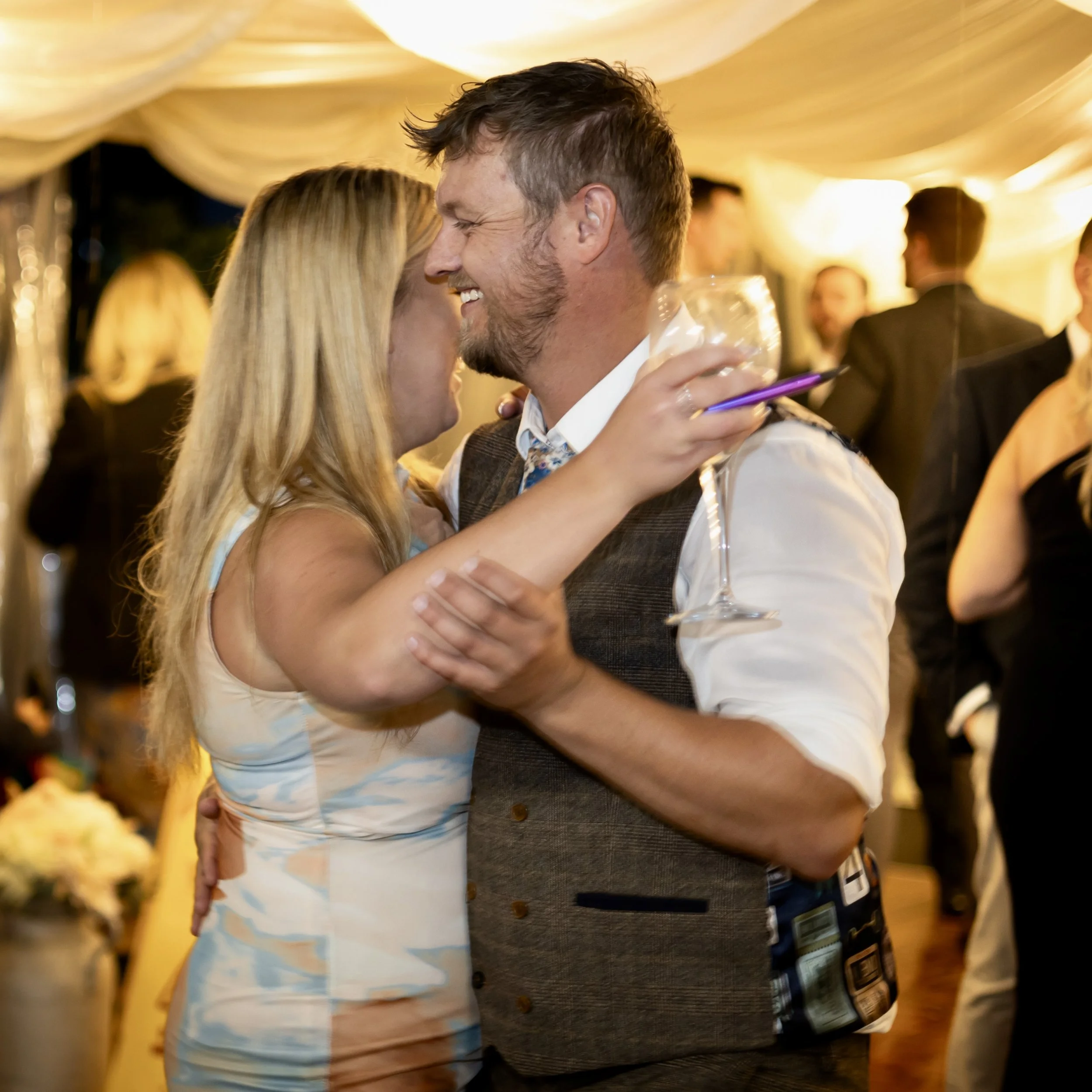 A man and woman embracing and smiling at each other at a celebration or wedding, in an indoor decorated venue.