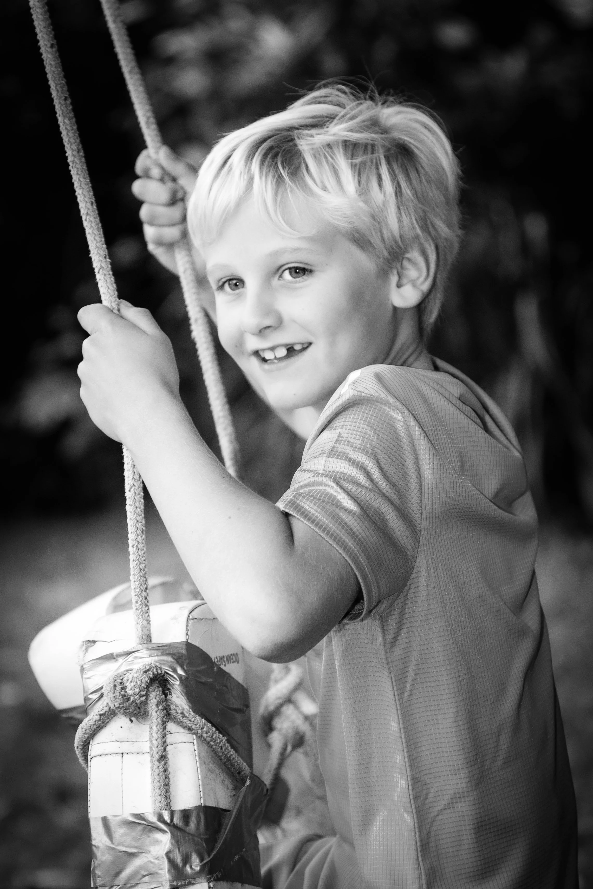 A young boy with light-colored hair smiling and holding onto a swing set chain in a blurry outdoor background.