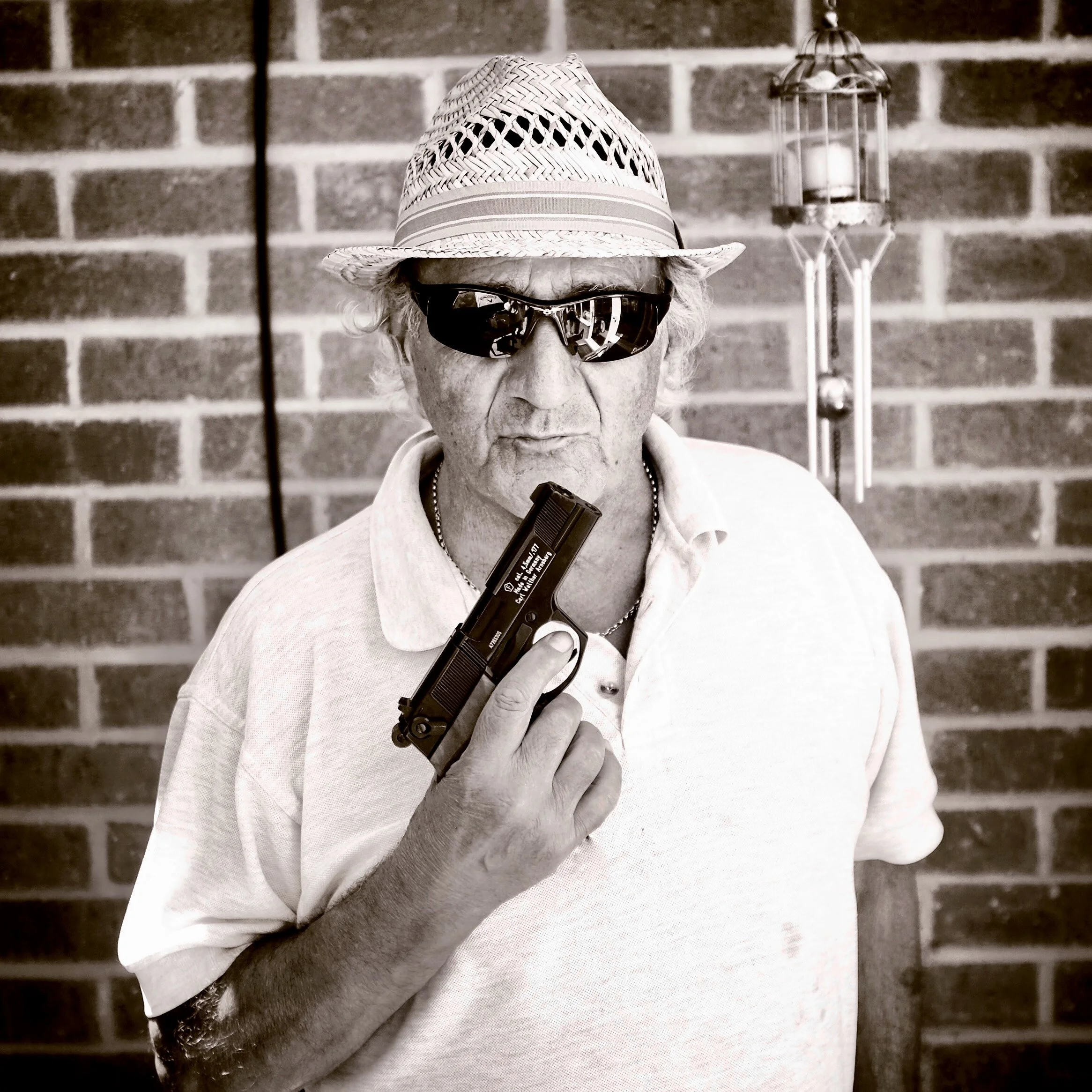 A person wearing a straw hat, sunglasses, and a white shirt, holding a gun against a brick wall background.