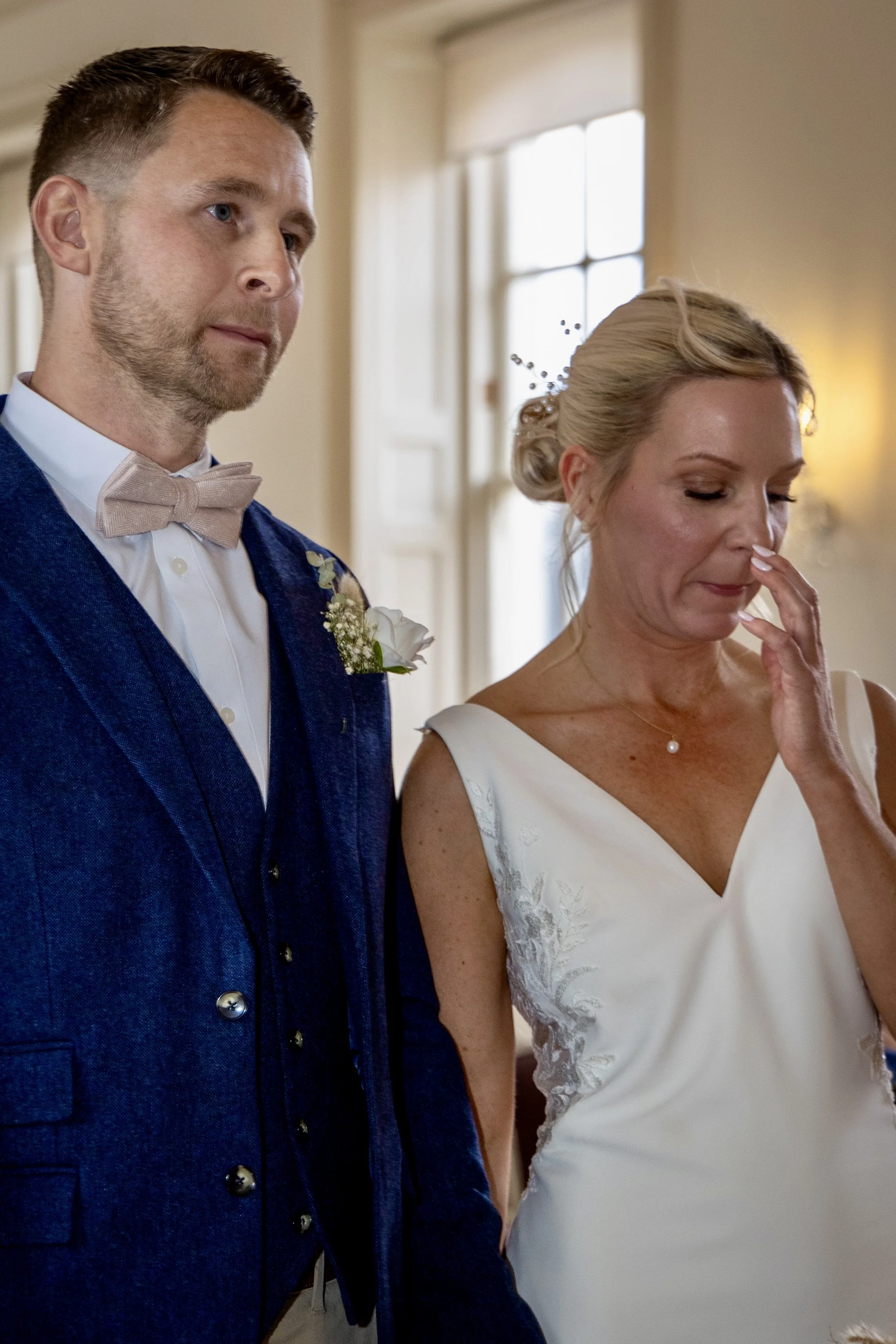 A man in a blue suit and beige bow tie standing beside a woman in a white dress in a room with beige walls and a window in the background.