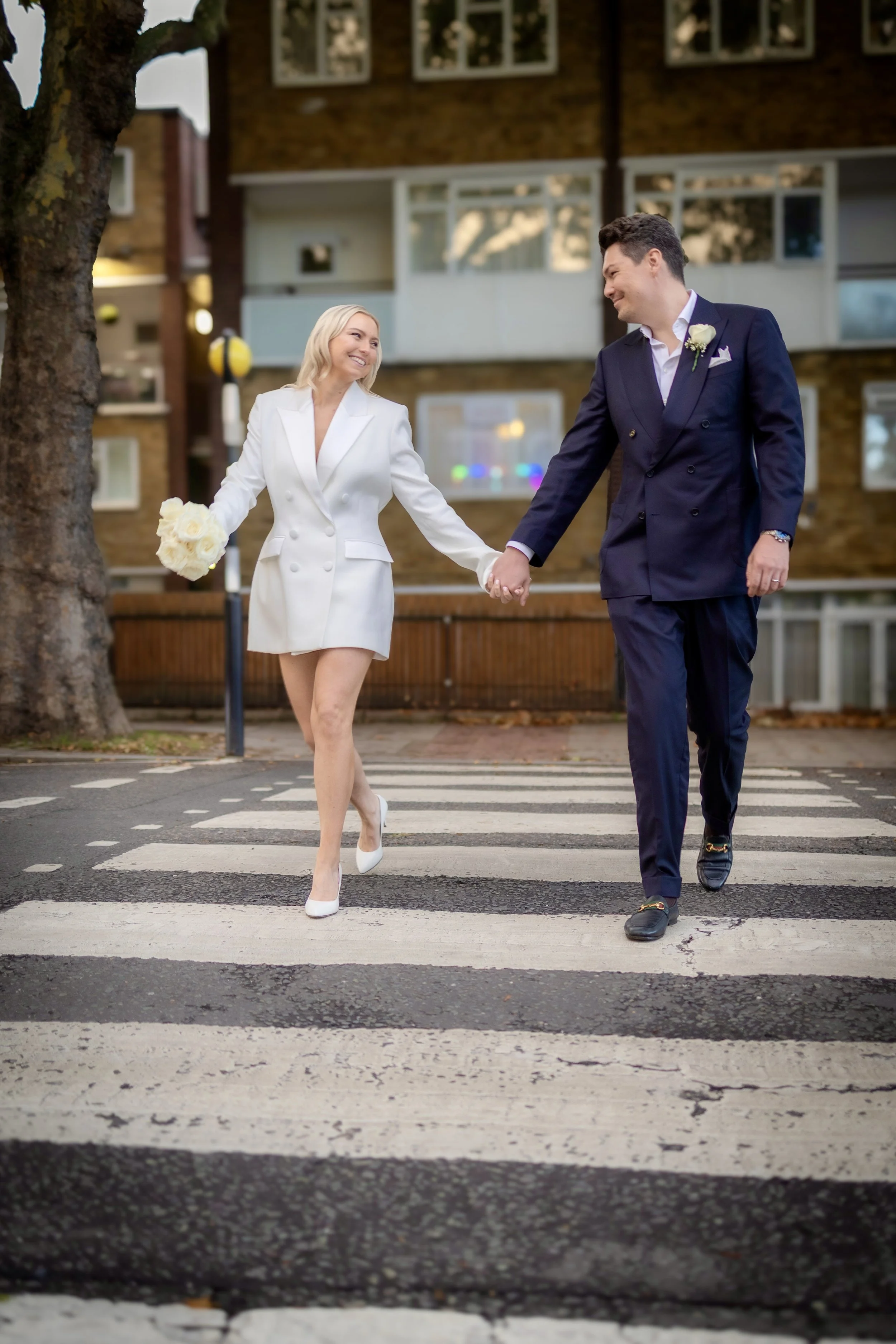 A happy couple in wedding attire walking hand-in-hand across a crosswalk in an urban setting.