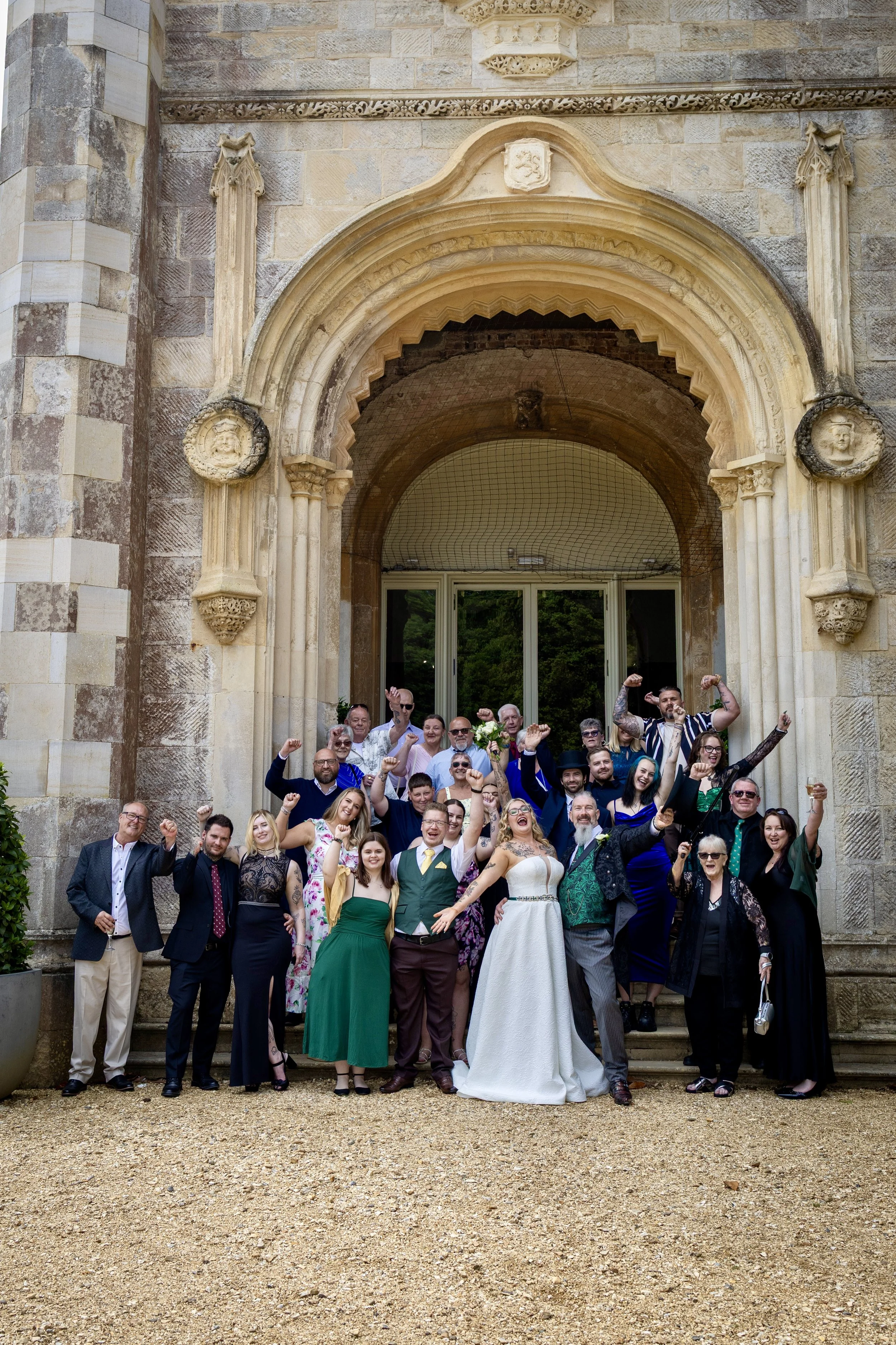 A large group of people gathered in front of an old stone building with a grand arched entrance, celebrating at a wedding. The bride in a white wedding gown and the groom in a dark suit are at the center, surrounded by family and friends in formal at