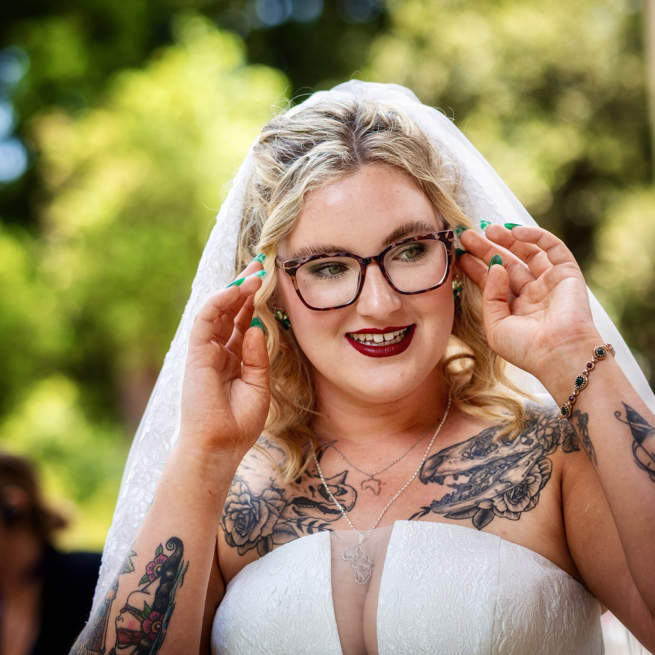 Close-up of a blonde woman in a white wedding dress adjusting her glasses, with tattoos on her arms, outdoors with green blurred background.