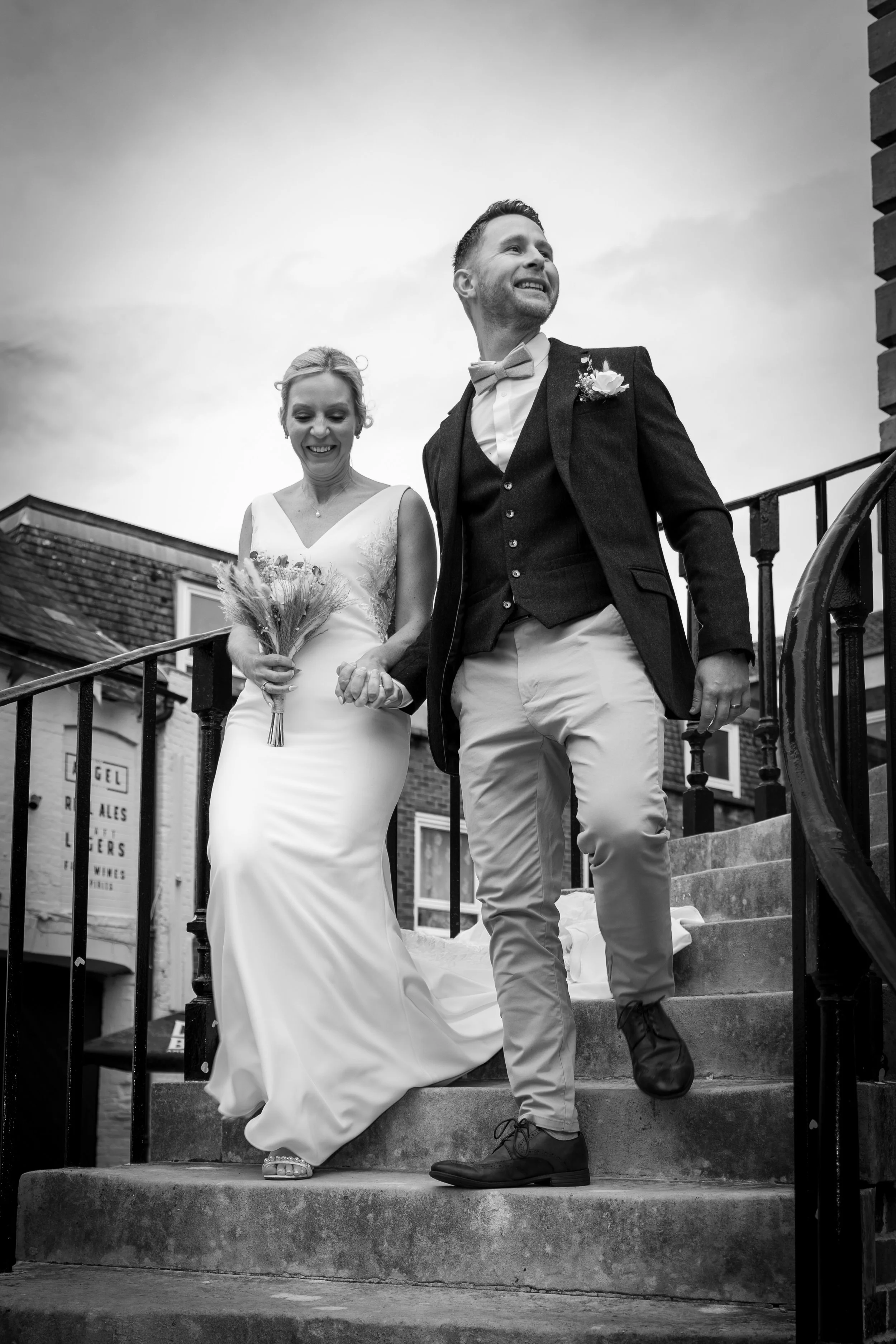 A bride and groom step down from stairs outside a building on their wedding day, holding hands and smiling.
