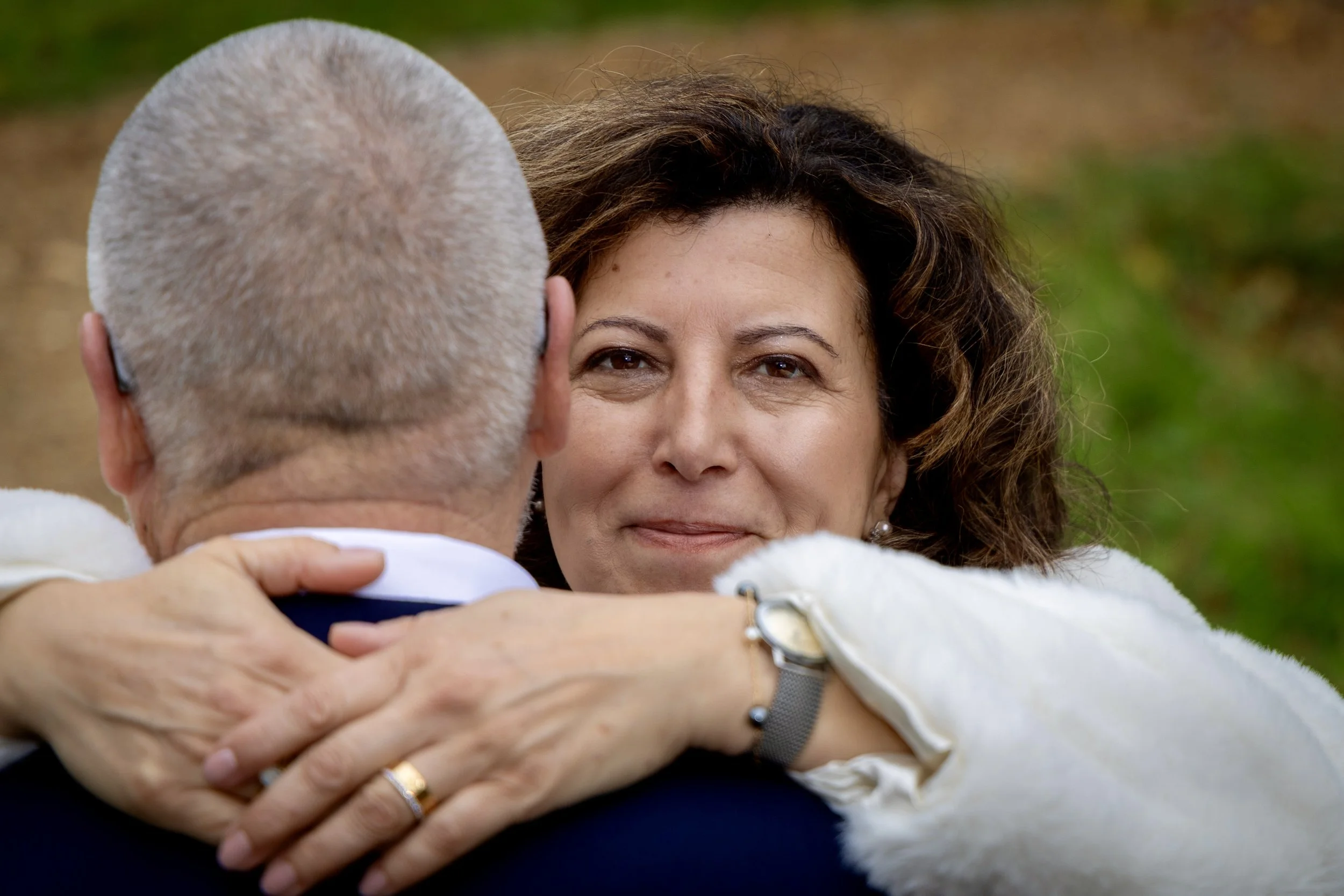 A woman with curly brown hair embracing an older man with a bald head outdoors, with a background of grass and trees.
