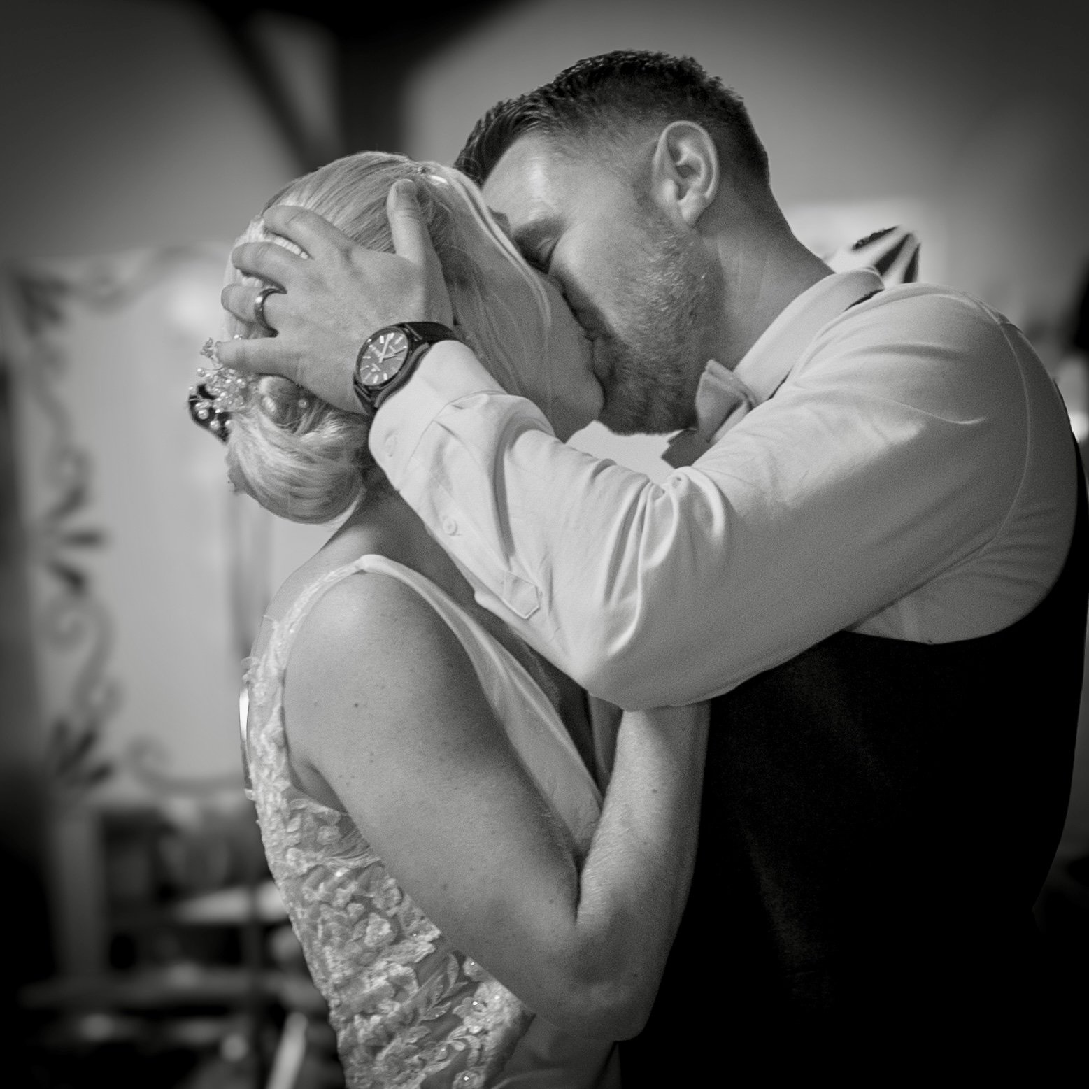 A black and white photo of a couple kissing, with the man holding the woman's head gently, their eyes closed. The woman has styled blonde hair and is wearing a detailed dress, while the man is dressed in a shirt and tie.