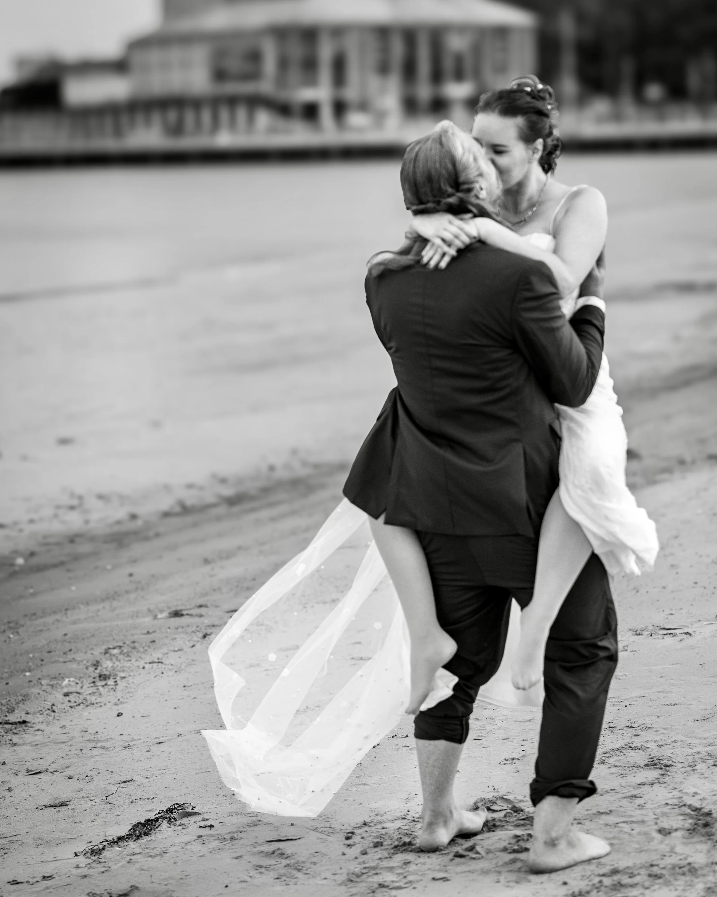 A man and woman embracing and kissing on the beach during the daytime, with a large building along the waterfront in the background. The woman is holding a piece of cloth or fabric that is flowing in the wind.