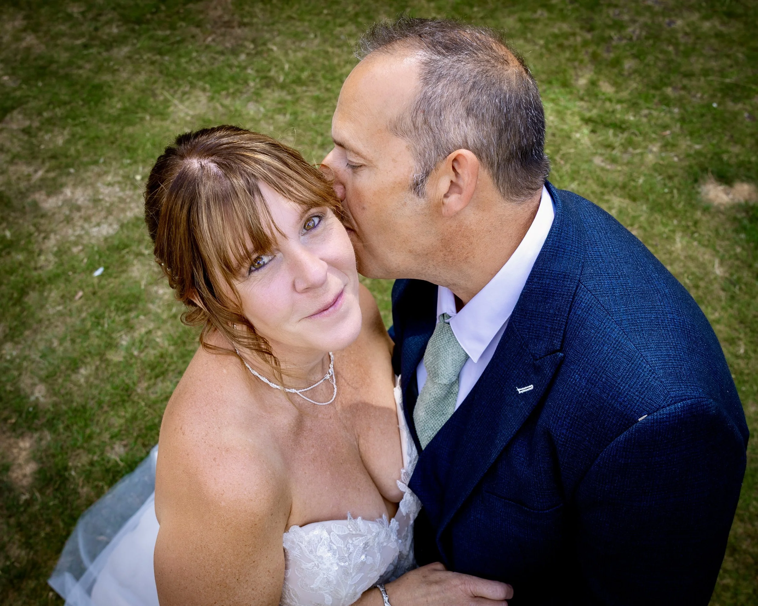 A bride and groom share a kiss outdoors on a grassy area, with the groom wearing a blue suit and the bride in a white dress with lace detail. The bride has red hair and light skin, and the groom has short, dark hair and light skin.