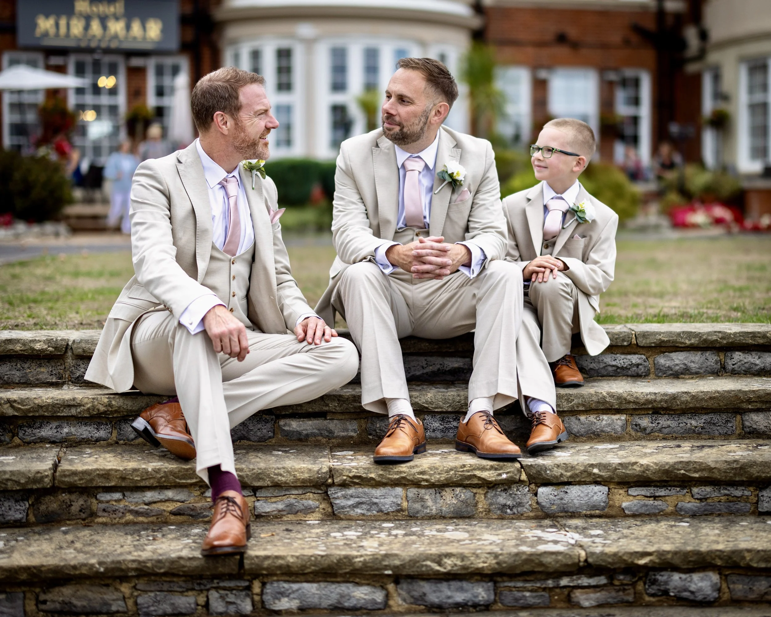 Three men and a boy dressed in beige suits sitting on stone steps outside a building having a conversation, with the building's exterior visible in the background.