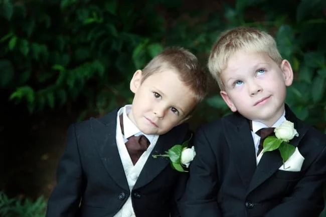 Two young boys in formal suits with boutonnières, sitting outdoors with greenery in the background, looking up and to the side.