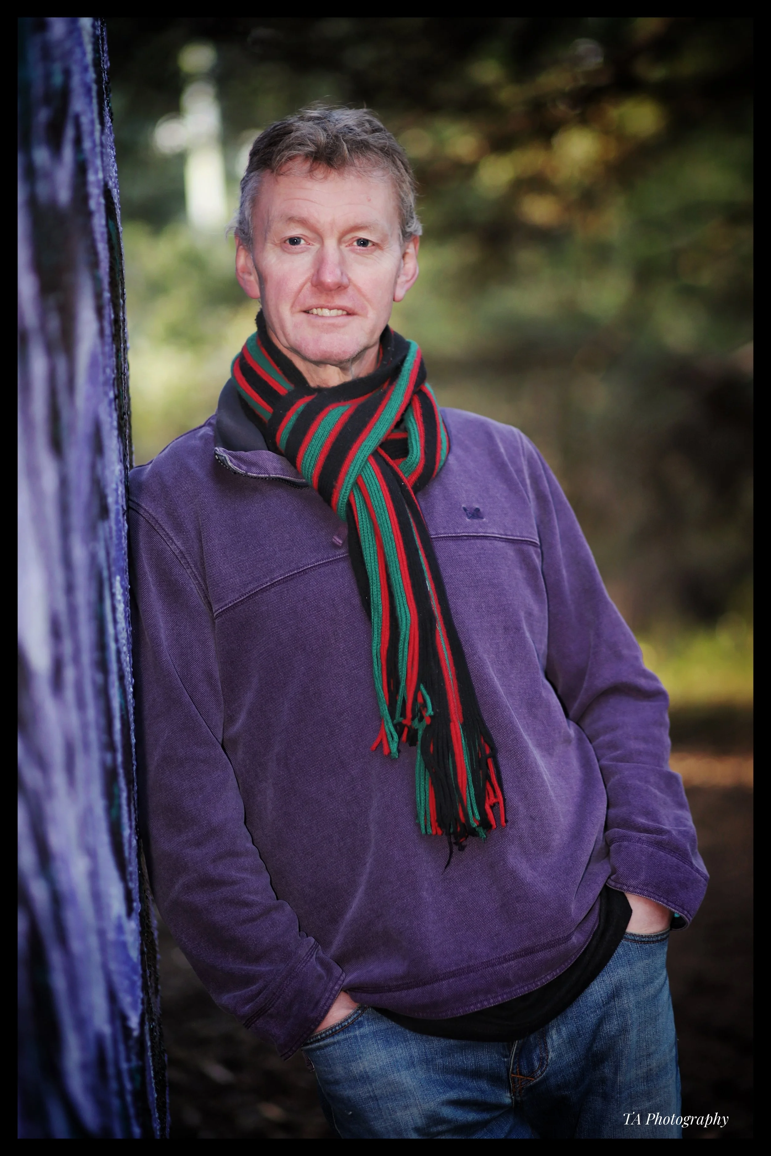 A man standing outdoors leaning against a purple painted wall with trees and blurred foliage in the background, wearing a purple jacket, a black hoodie, and a colorful striped scarf.
