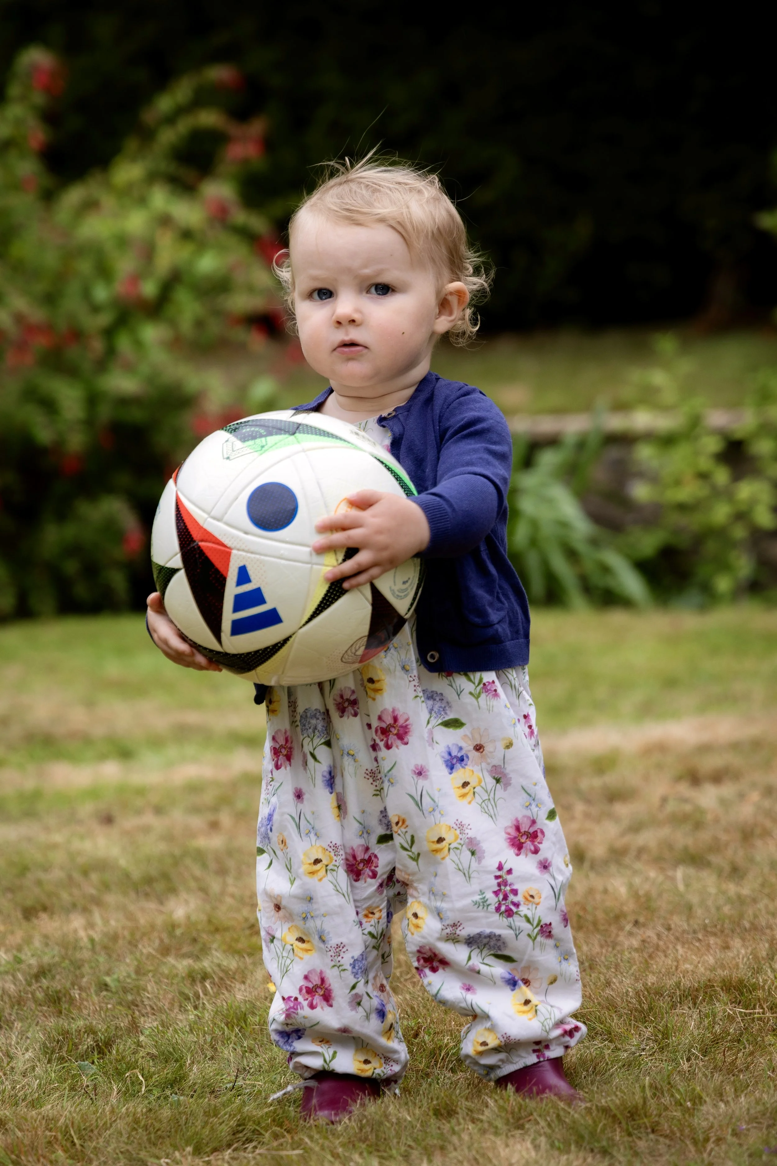 A young girl with blonde hair and blue eyes stands outdoors, holding a soccer ball, wearing a floral jumpsuit, a blue jacket, and purple rain boots, with a blurred green leafy background.
