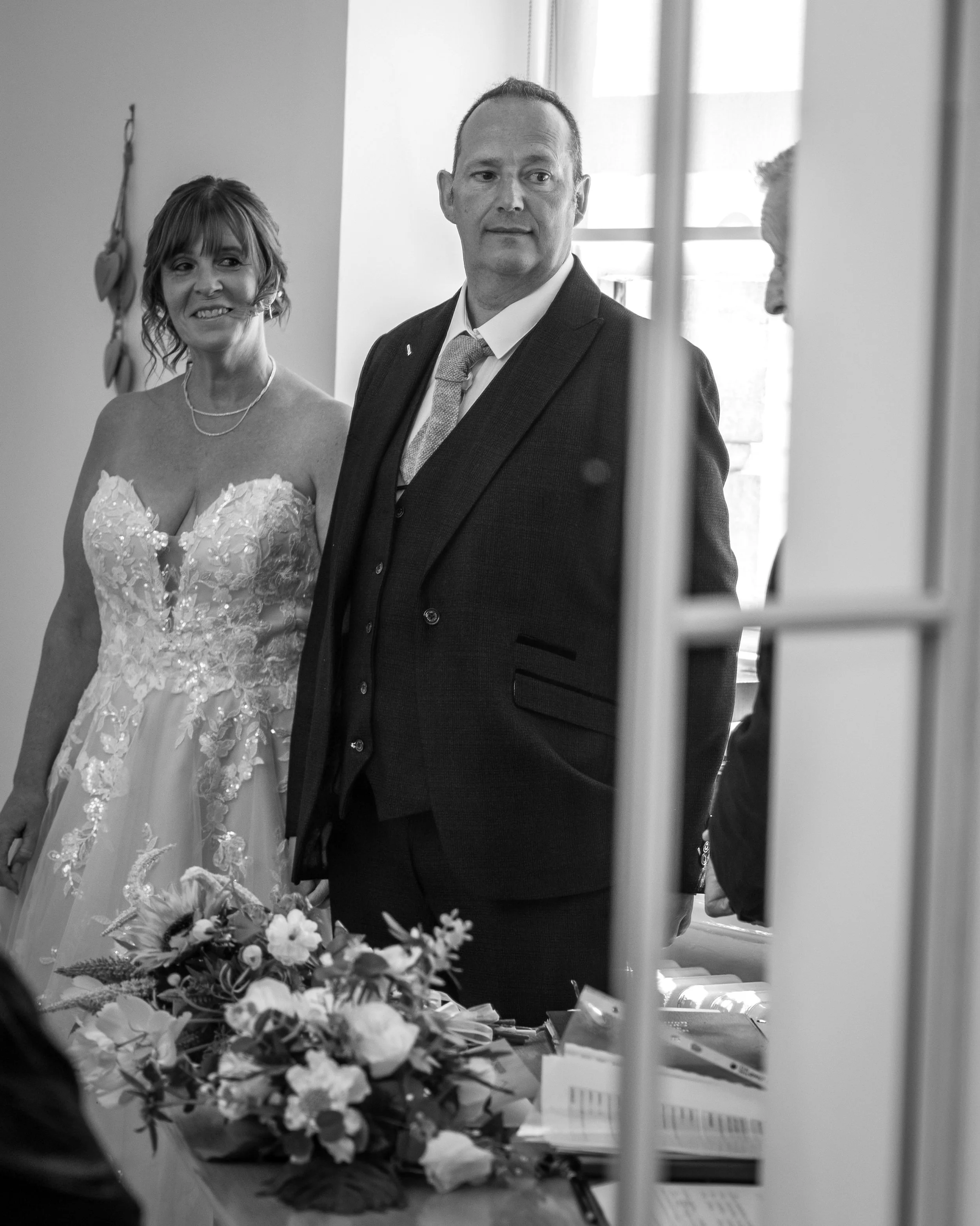 Black and white photo of a man and woman standing side by side, dressed formally. The woman is in a wedding dress, and they are inside a room with a window in the background. There are flowers on a table in the foreground.