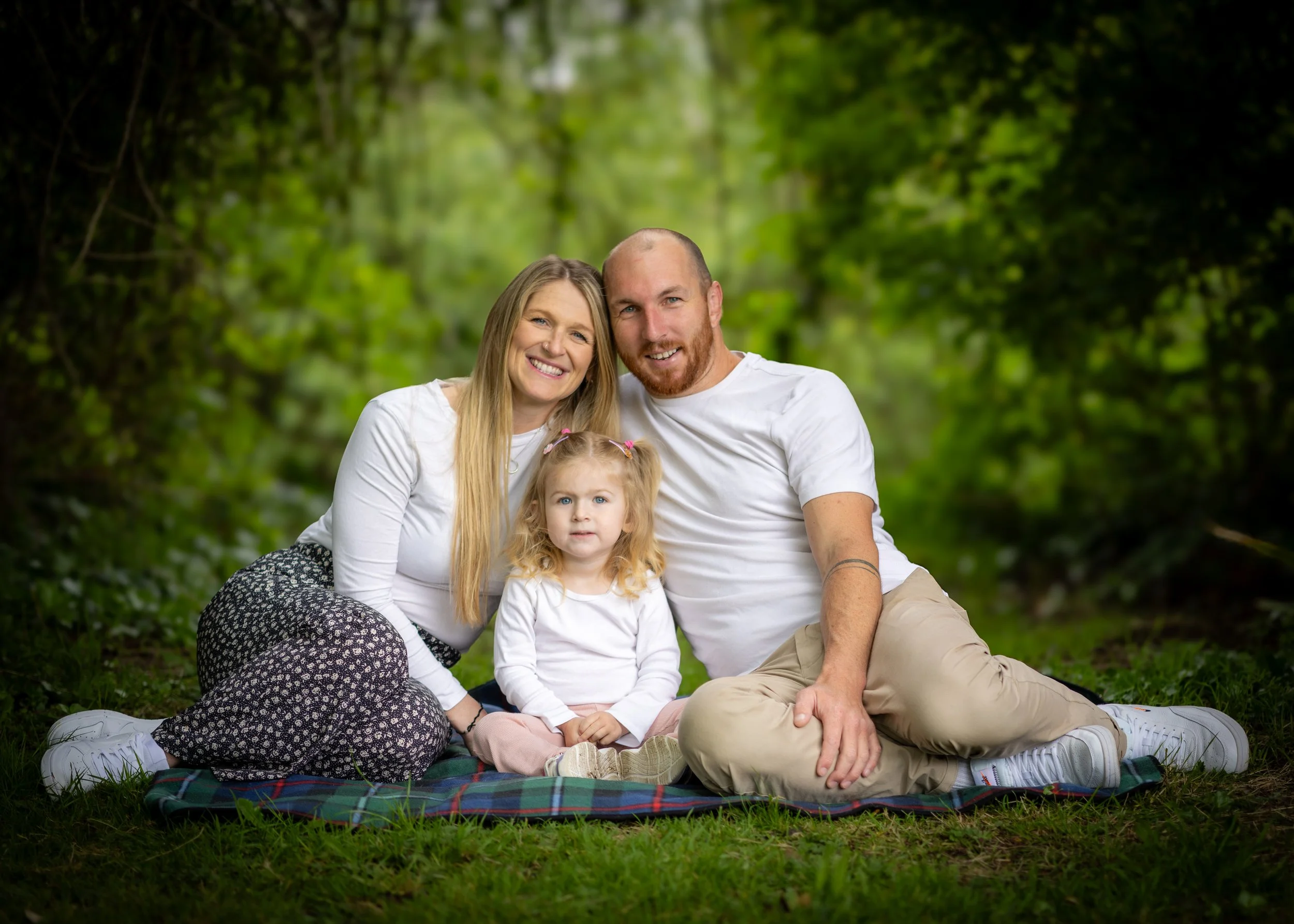 A happy family of four sitting on a blanket in a forested park, smiling and enjoying a sunny day. The family includes a woman, a man, a young girl, and a toddler girl.