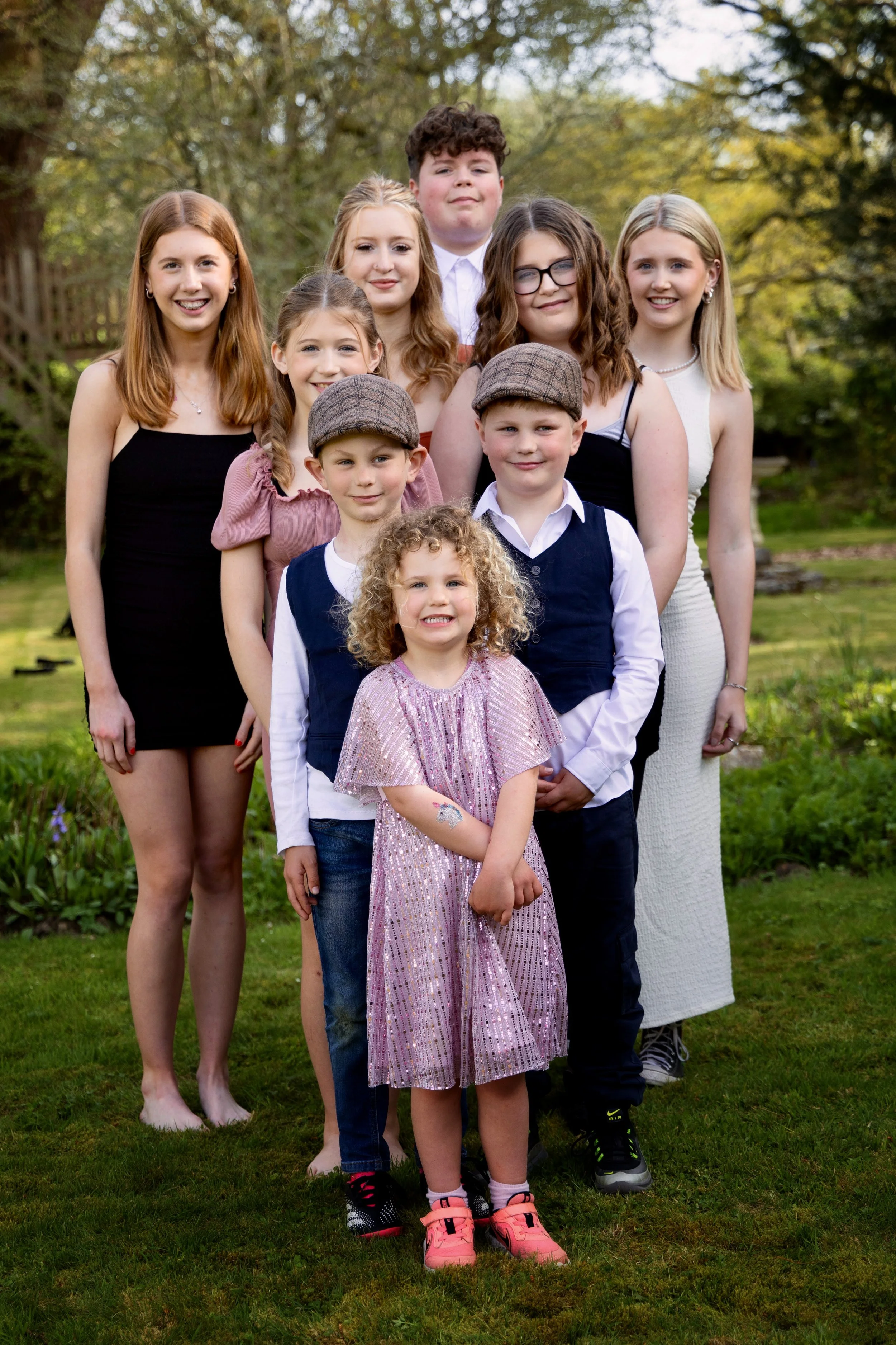Group of children and teenagers standing outdoors on grass during the day, smiling for the photo with trees in the background.