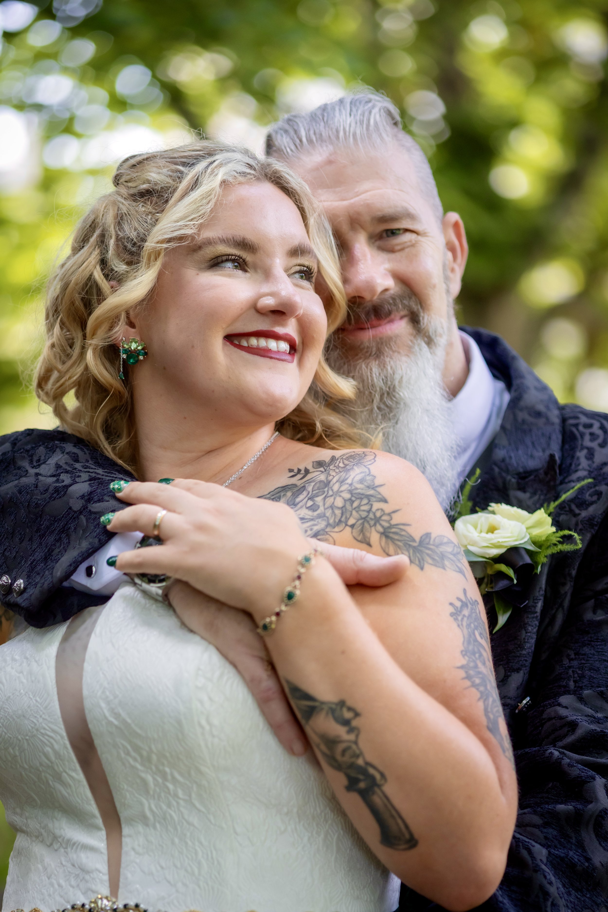 Close-up photo of a smiling woman with blonde hair and a tattoo on her arm, and a man with gray hair and a beard, both looking happy, outdoors with blurred green trees in the background, appearing to be a couple or friends.