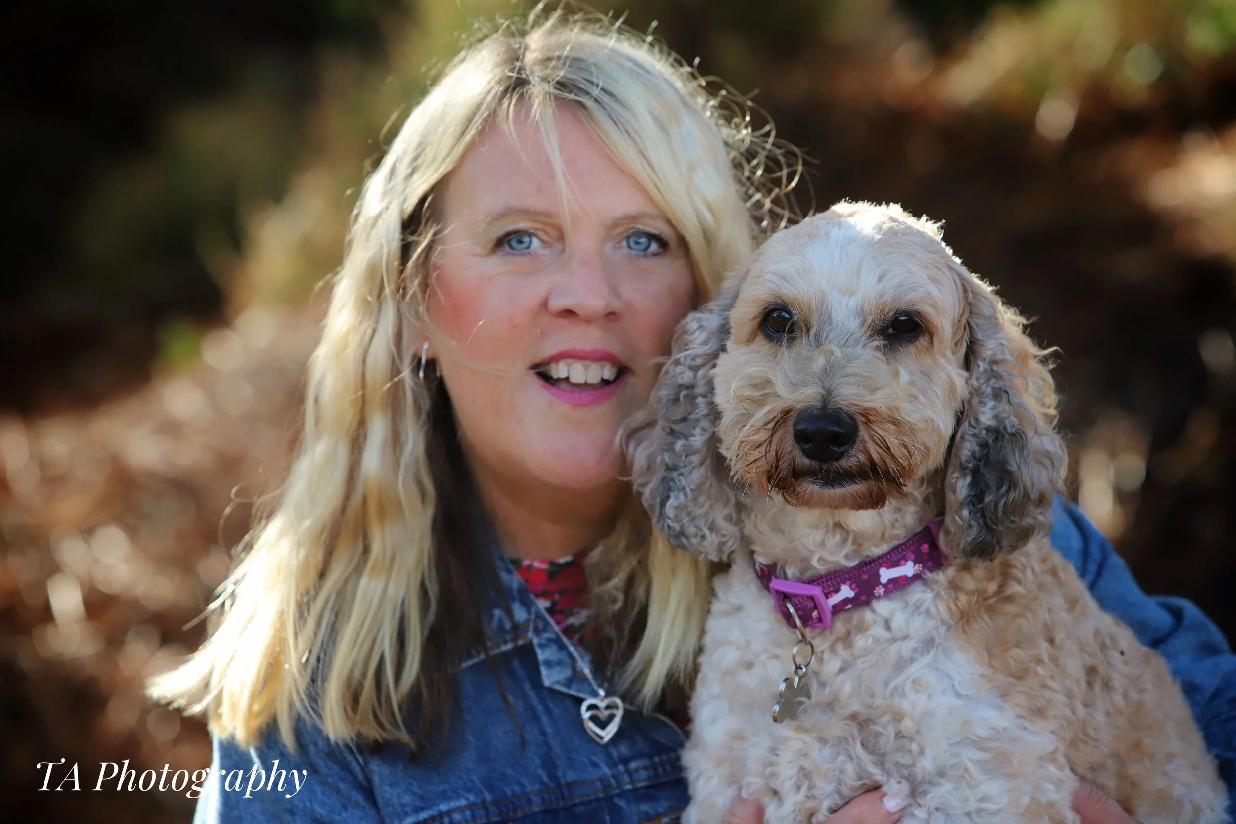 A woman with long blonde hair and blue eyes holding a curly-haired beige dog wearing a pink harness outdoors with blurred fall foliage in the background.