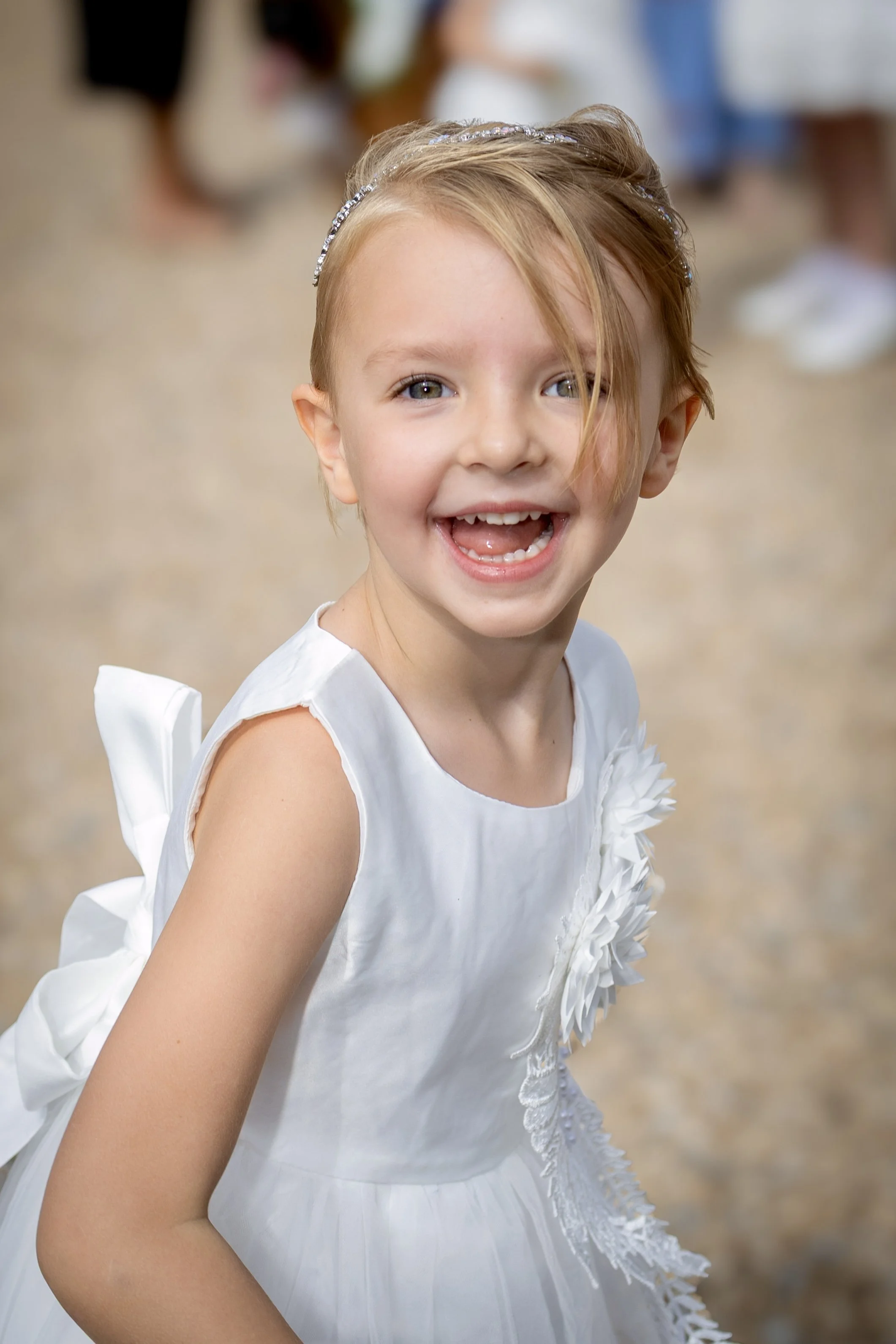 A young girl with light skin, blond hair, blue eyes, wearing a white dress with floral decorations, smiling and showing her teeth.