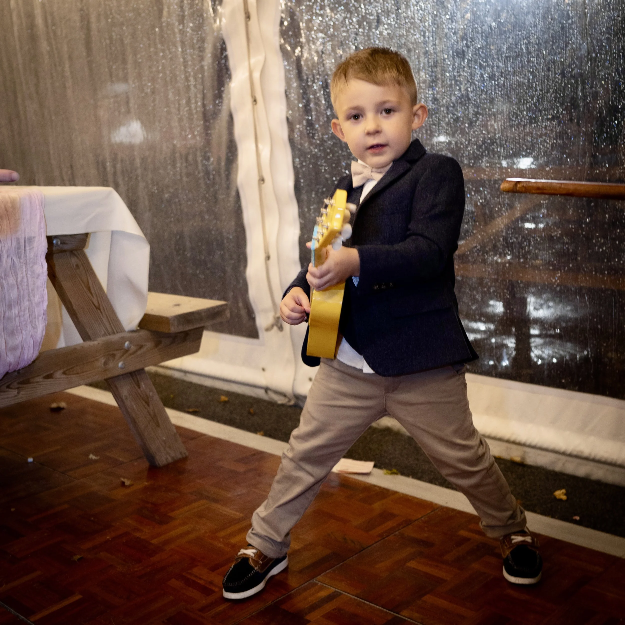 A young boy with short brown hair wearing a black blazer, white shirt, beige pants, and brown shoes standing with his legs apart, holding a yellow toy guitar, on a wooden floor in front of a sparkly silver backdrop.