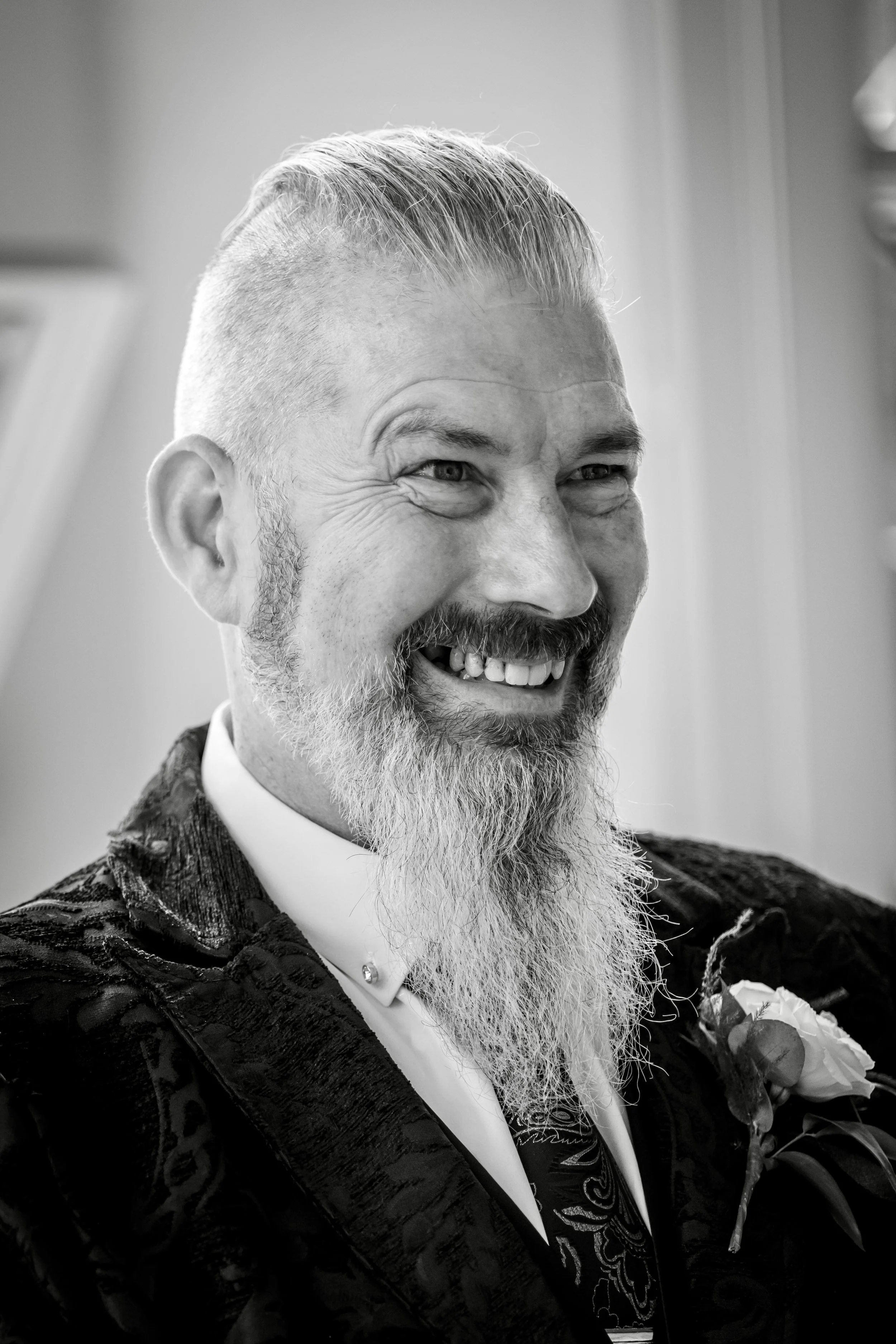 A close-up black and white photo of a middle-aged man with a beard and mustache, smiling and wearing a patterned jacket with a boutonnière, likely at a formal event.