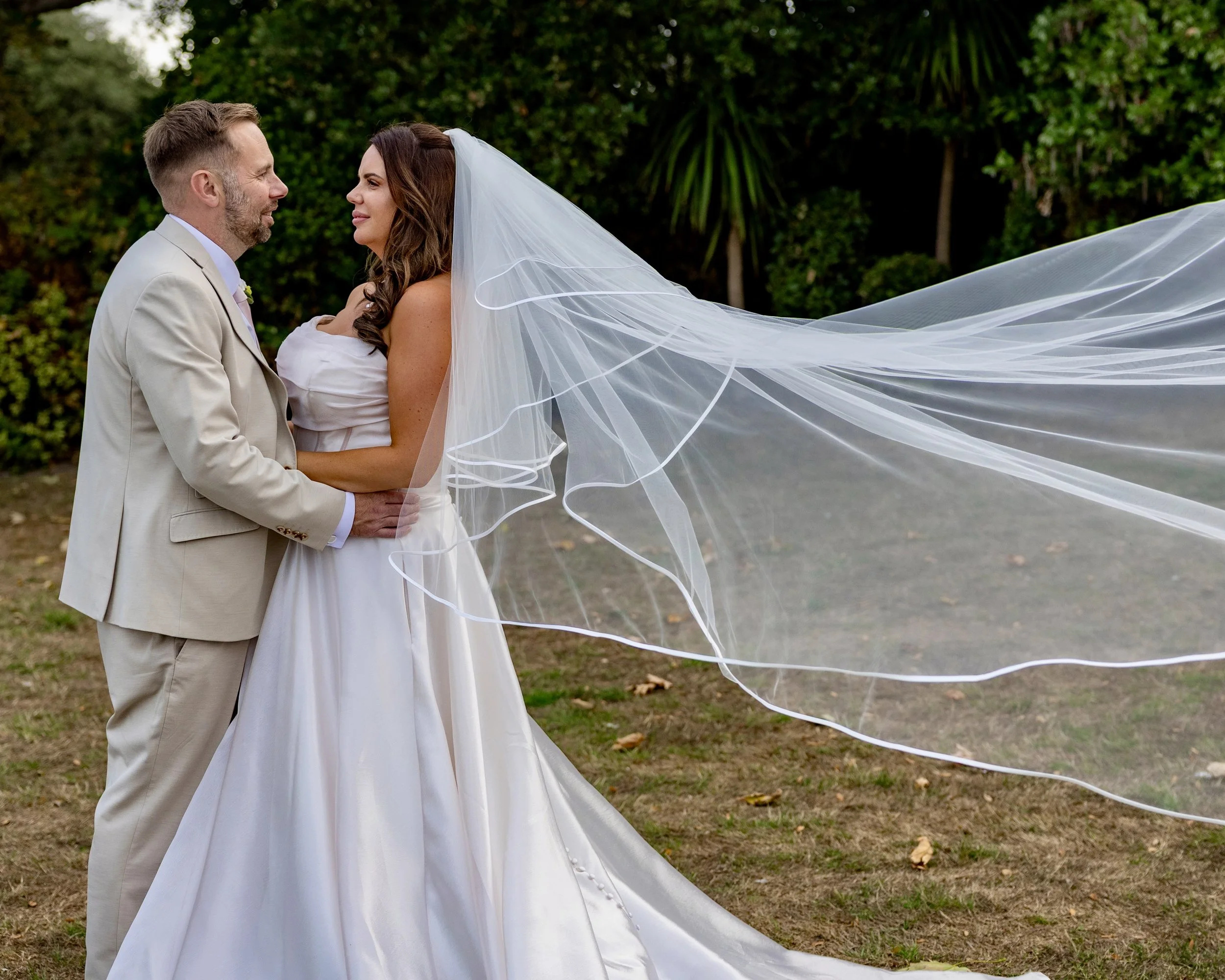 A groom and bride are facing each other outdoors, with the groom wearing a beige suit and the bride in a white wedding dress and veil. They are smiling and holding each other's hands, with a backdrop of green trees and bushes.