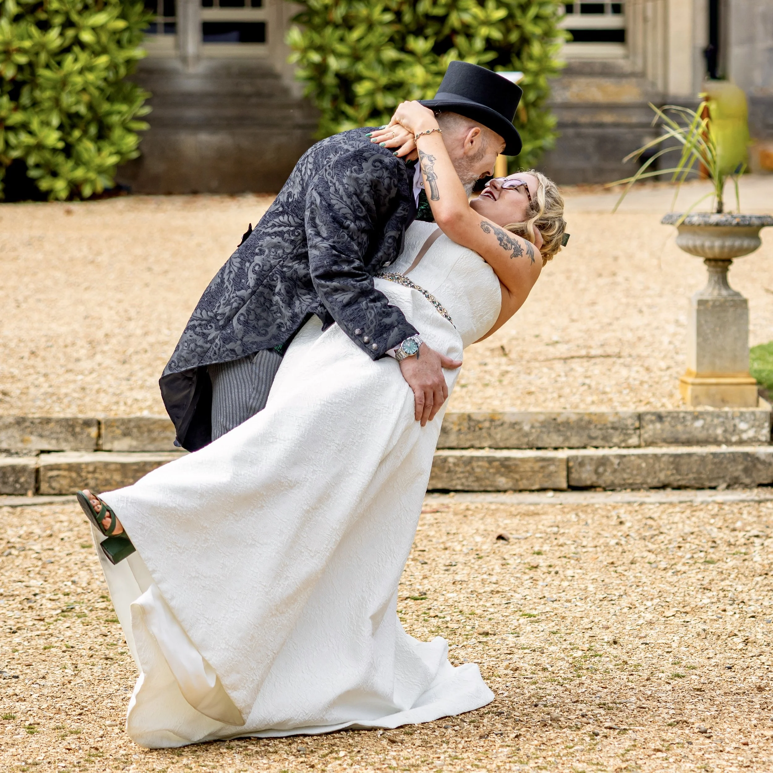 A couple dressed in wedding attire sharing a dance outdoors, with the groom wearing a black top hat and the bride in glasses and a white dress, surrounded by greenery and stone steps.