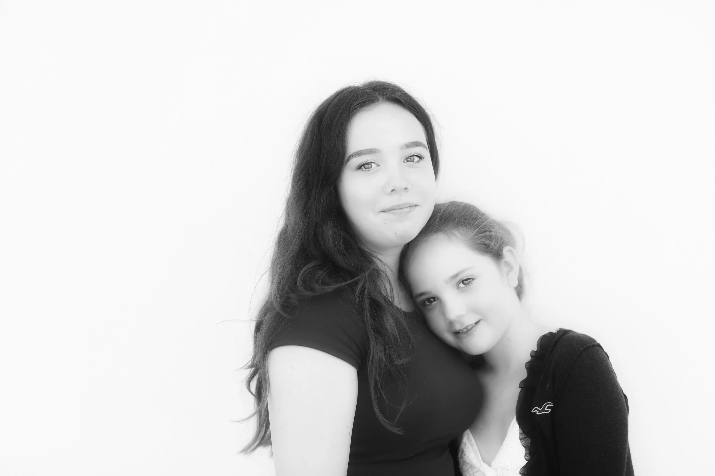A young woman with long, dark hair holding a smiling young girl with light hair close to her chest. They are standing against a plain white background.