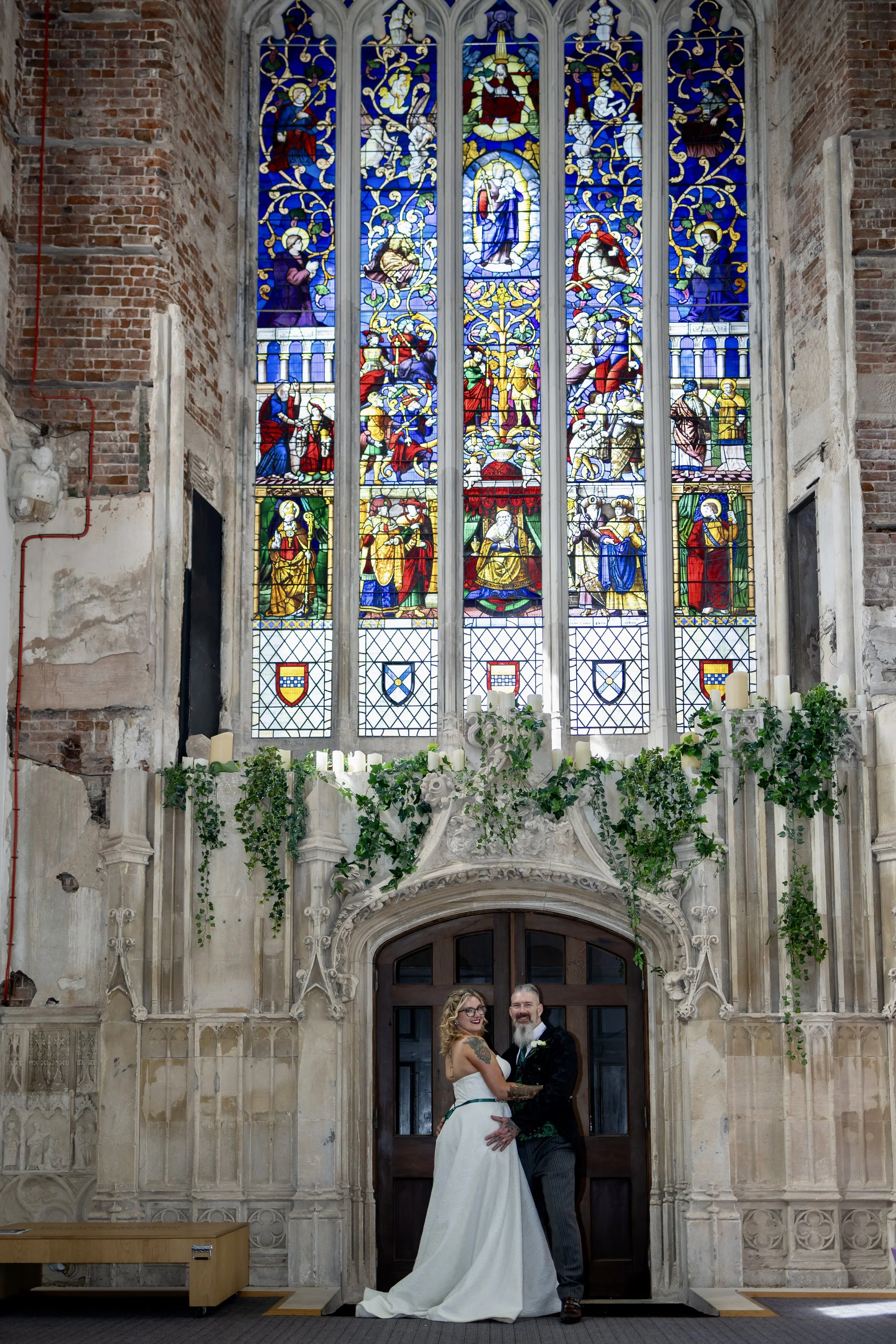 A bride and groom standing together inside a church with stained glass windows behind them. The bride is in a white wedding dress, and the groom is in a dark suit with a boutonniere. They are smiling and holding each other.