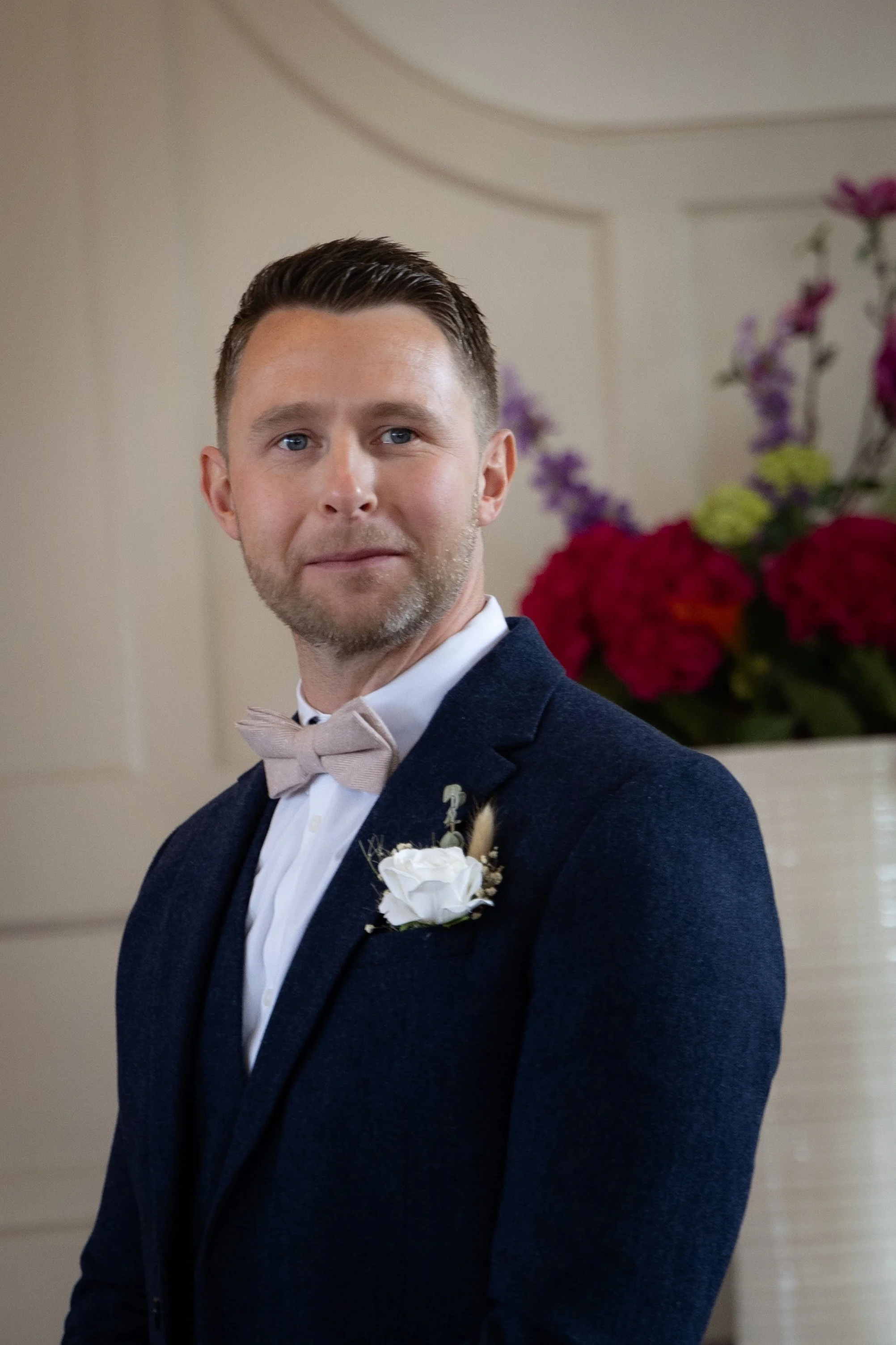 A groom in a dark blue suit with a light beige bow tie and a white boutonnière, standing indoors with a floral arrangement in the background.