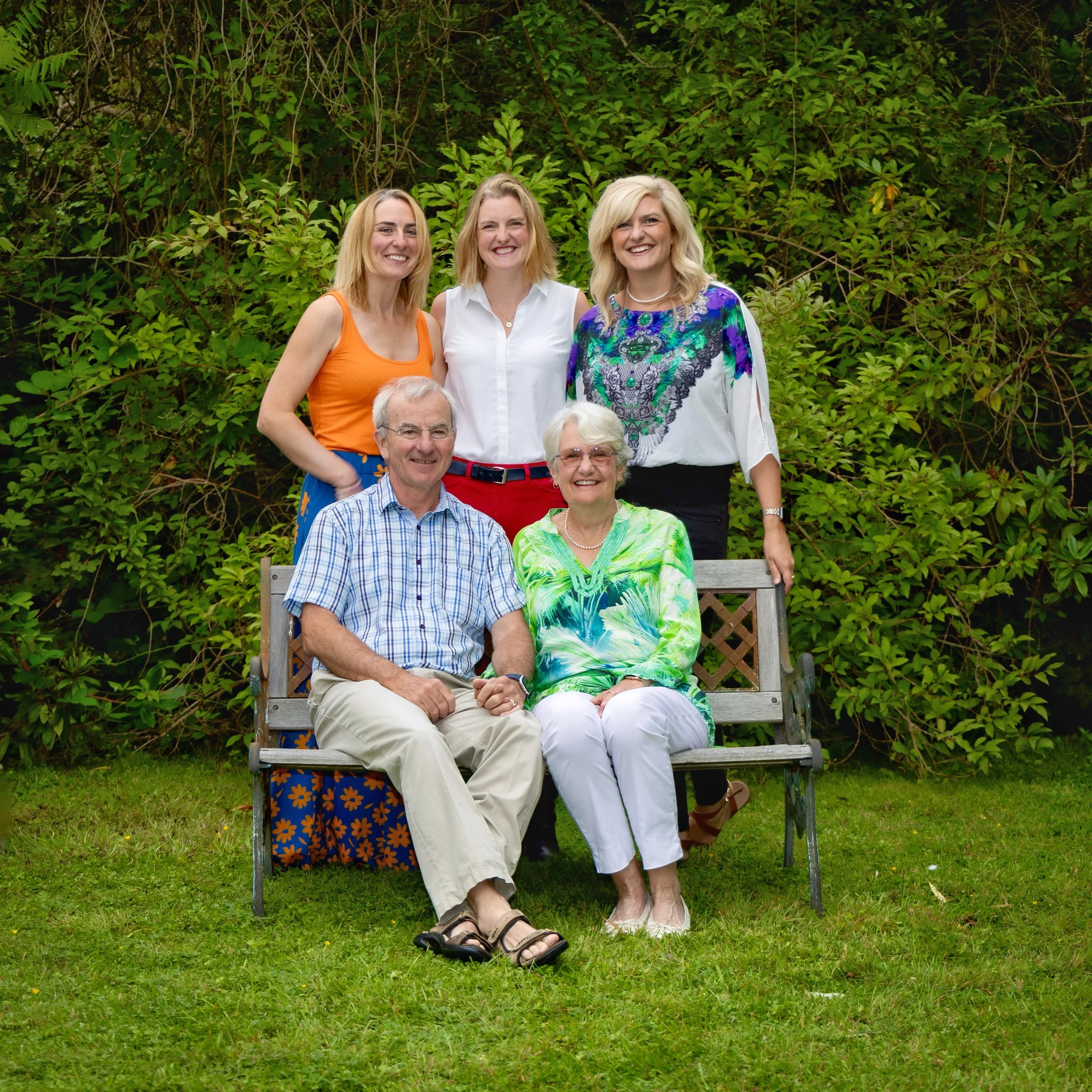 A family of six people posing together outdoors on a grassy area, with a backdrop of green foliage. The group includes three women with blonde hair standing behind, and an elderly man with gray hair and a woman with white hair sitting on a bench in the front.