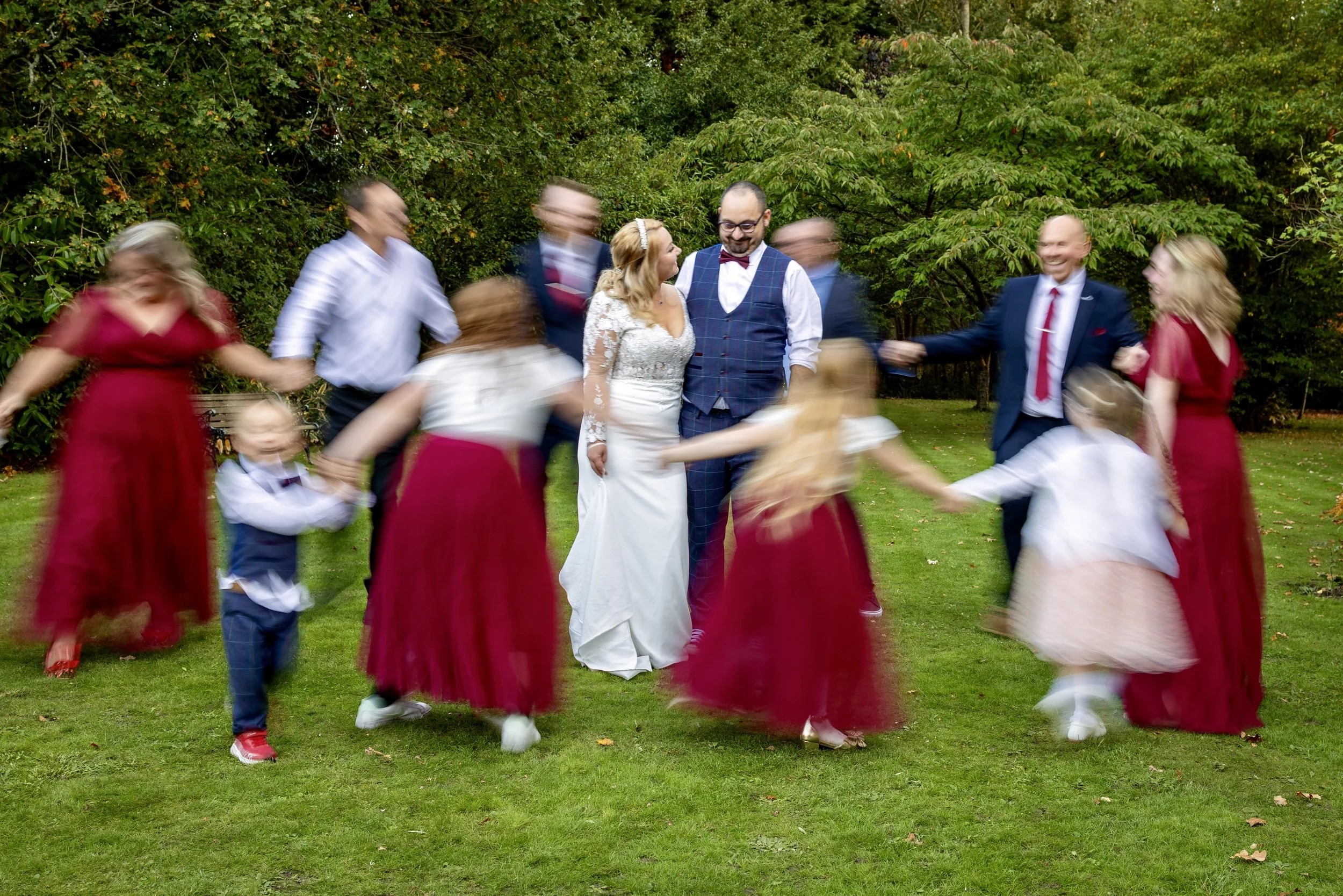 A wedding party dancing outdoors on a grassy area with trees in the background, with some people blurred from motion.