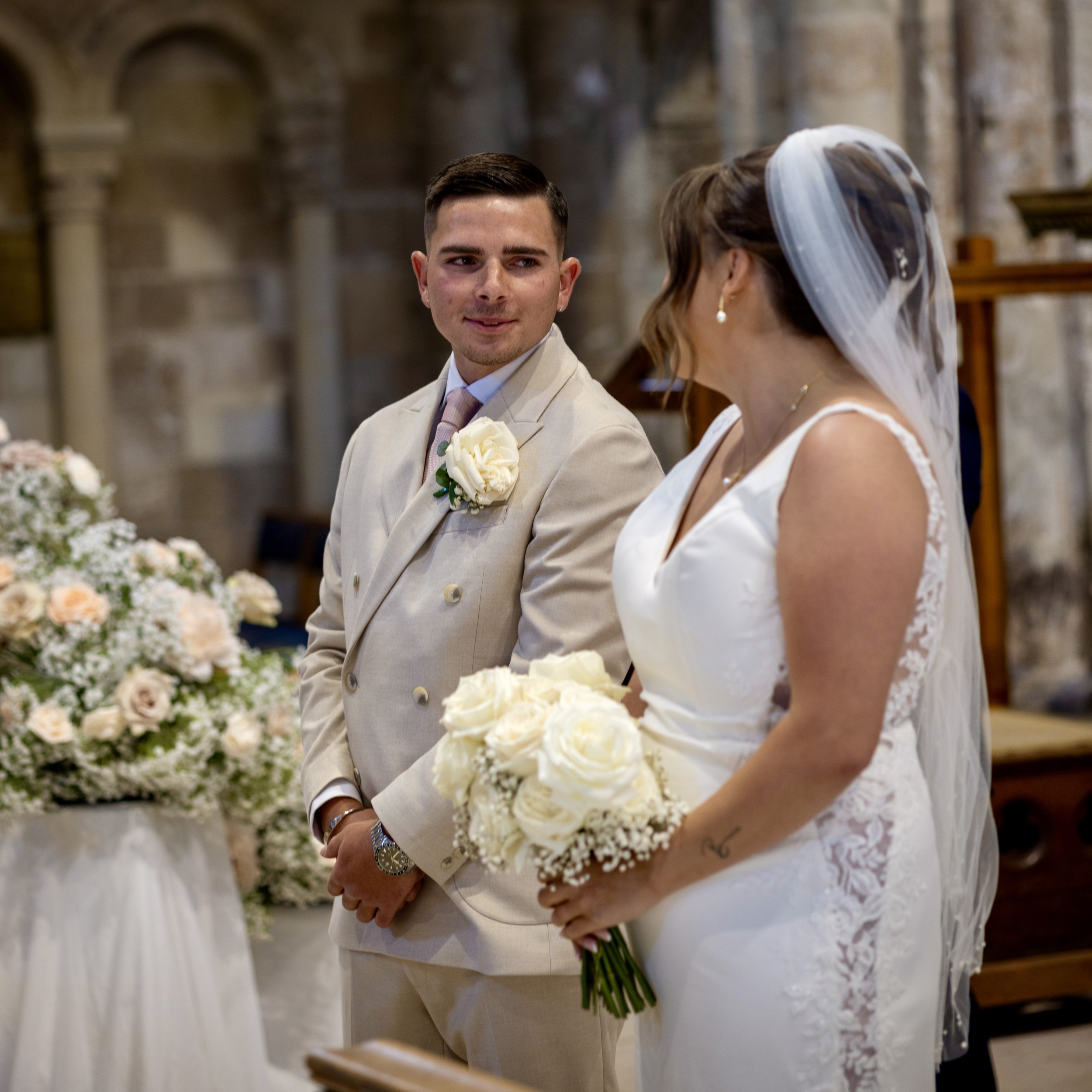 A bride and groom at their wedding ceremony inside a church, exchanging vows and holding bouquets of white roses.
