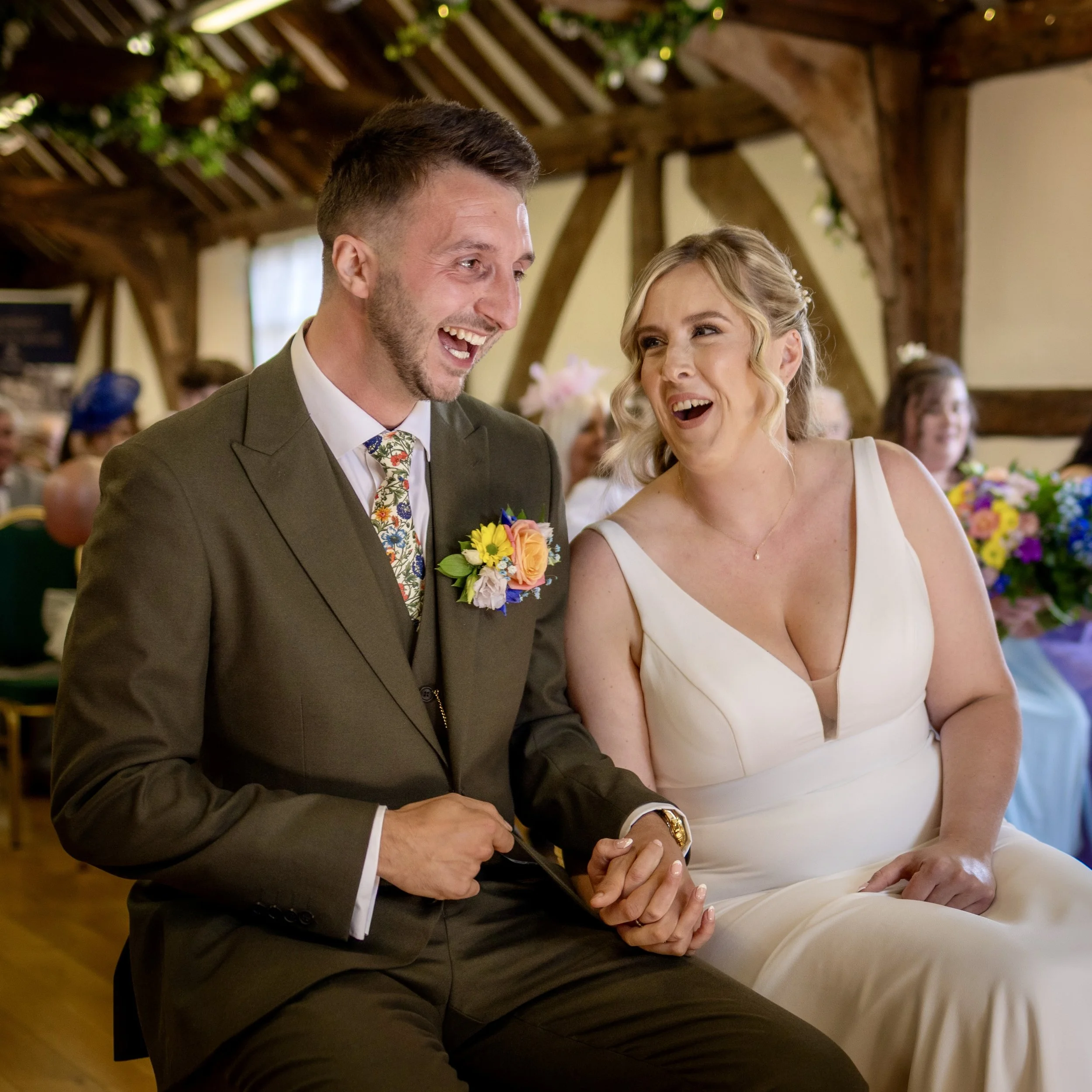 A man and a woman seated at a wedding reception, smiling and holding hands, with a decorated rustic wooden ceiling and floral arrangements in the background.