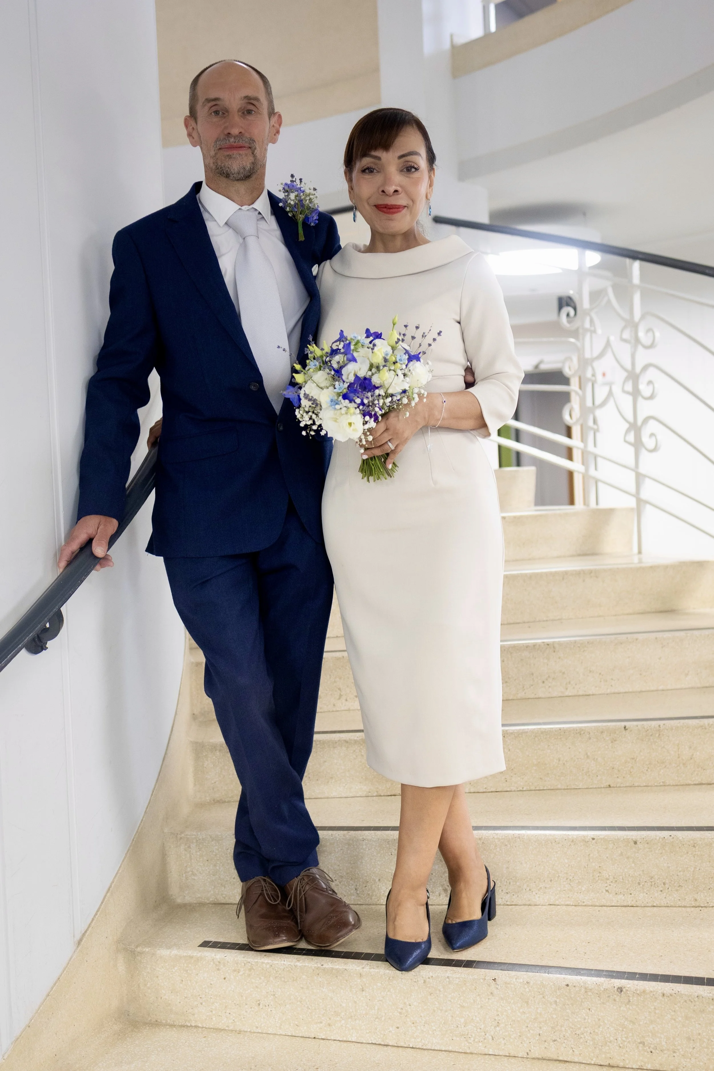 A couple dressed in wedding attire standing on staircase, with the man in a blue suit and white tie, and the woman in a white dress holding a bouquet of flowers, smiling at the camera.
