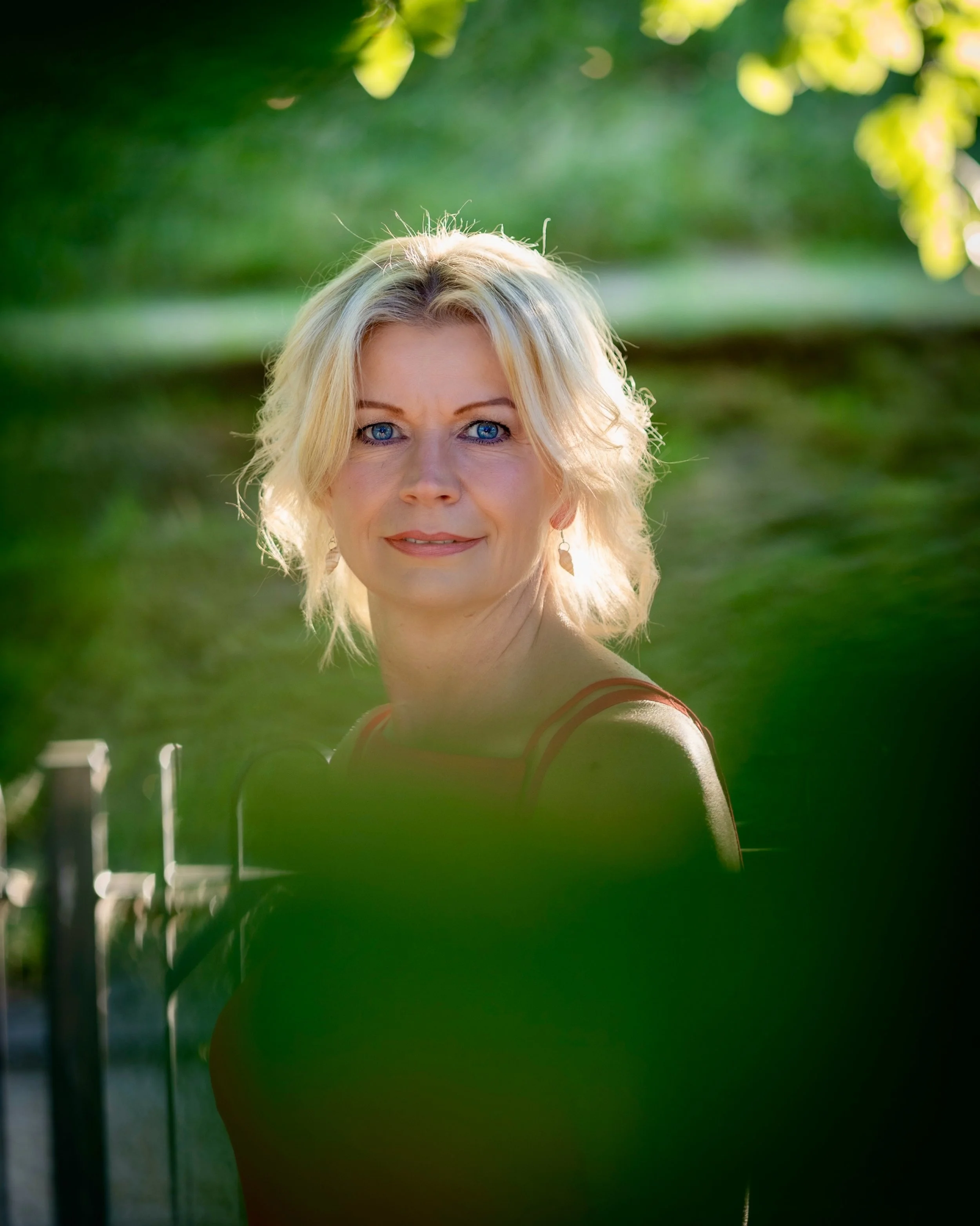 A portrait of a woman with blonde hair and blue eyes standing outdoors, with greenery and trees in the background, sunlit during daytime.