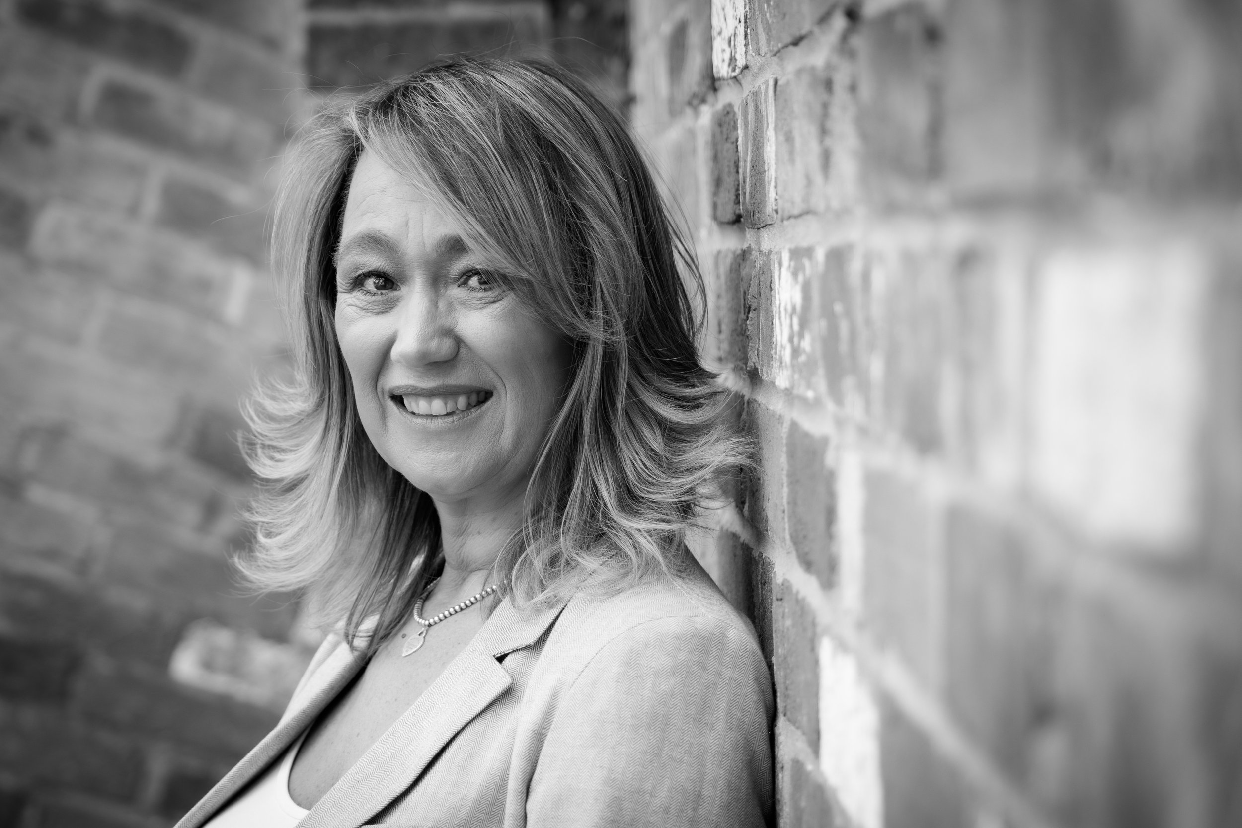 Black and white portrait of a woman with shoulder-length hair, smiling, wearing a blazer and a necklace, leaning against a brick wall.