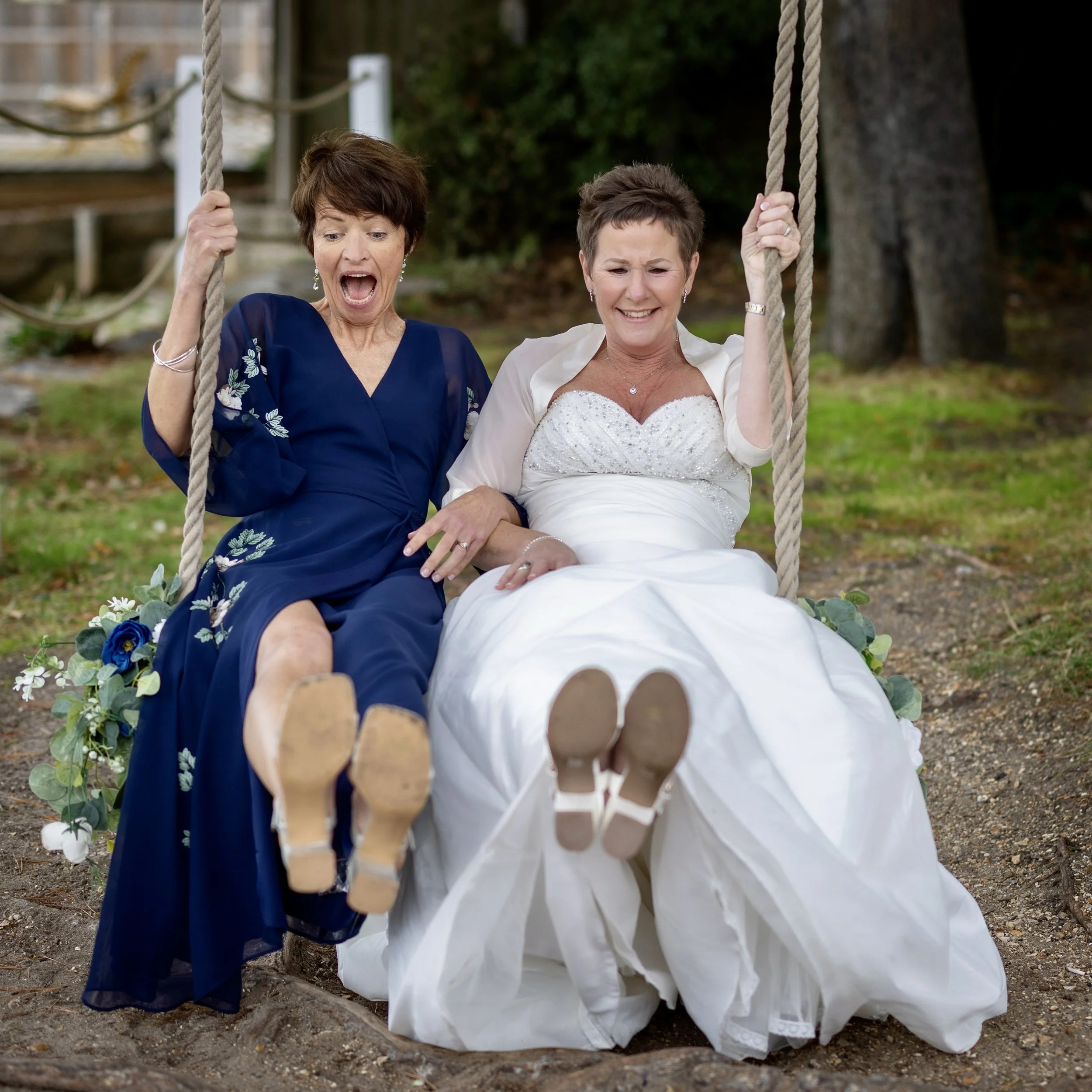 Two women, one in a wedding dress and one in a navy blue dress, sitting on a swing outdoors, smiling and enjoying each other's company.