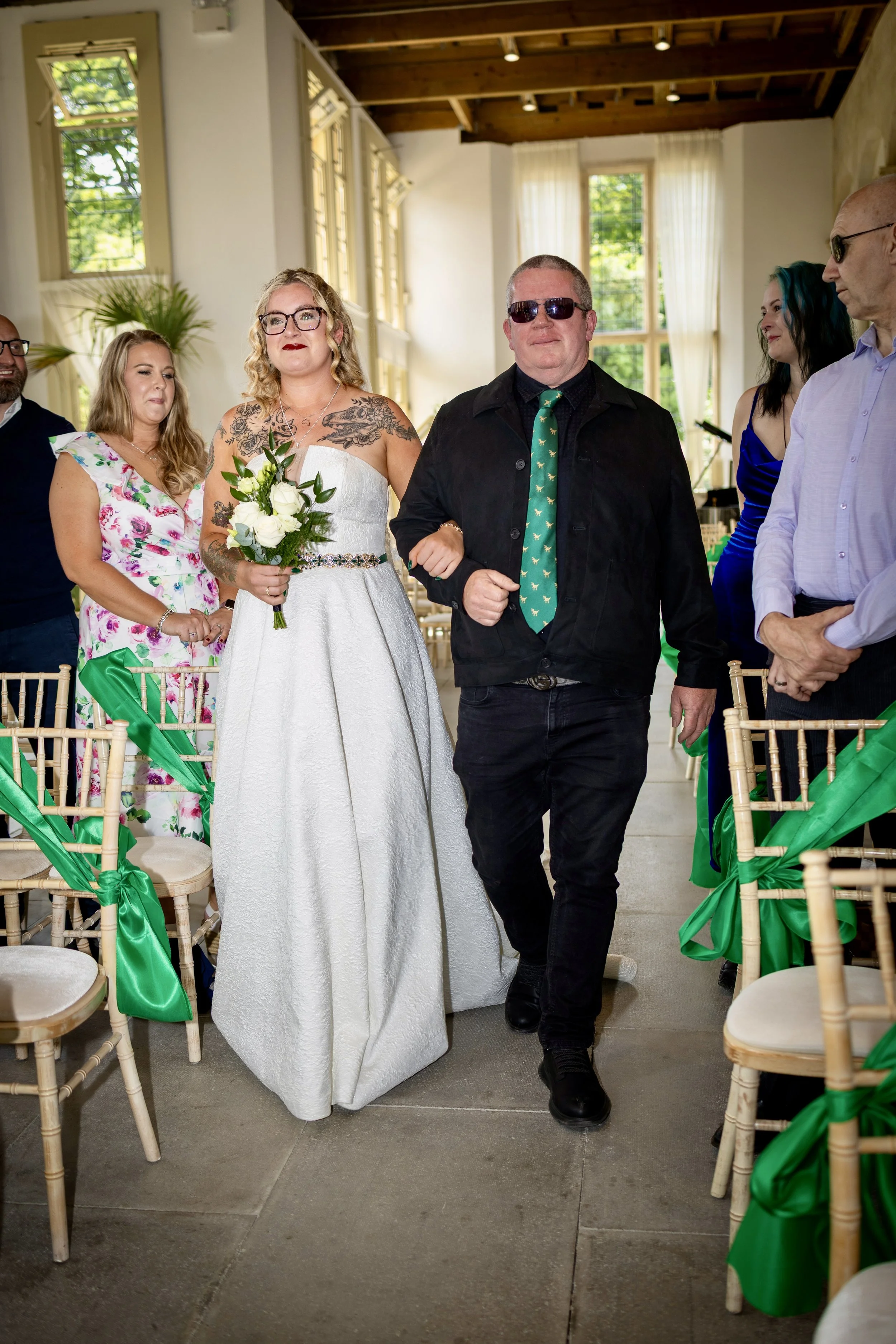 A woman in a white wedding dress holding a bouquet and walking down an aisle lined with chairs decorated with green satin bows, in a bright room with large windows and wooden ceiling beams.