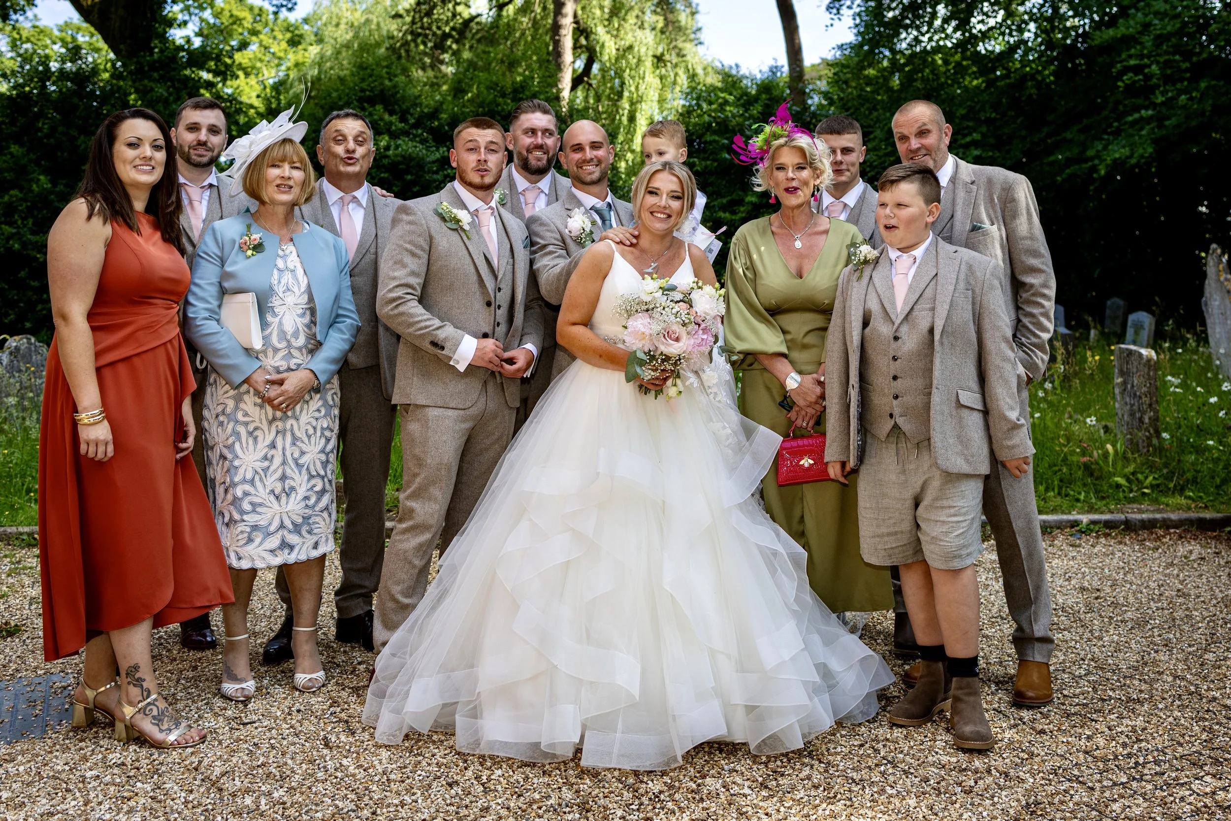 A wedding group photo outdoors with the bride in a white gown holding a bouquet surrounded by family and friends.