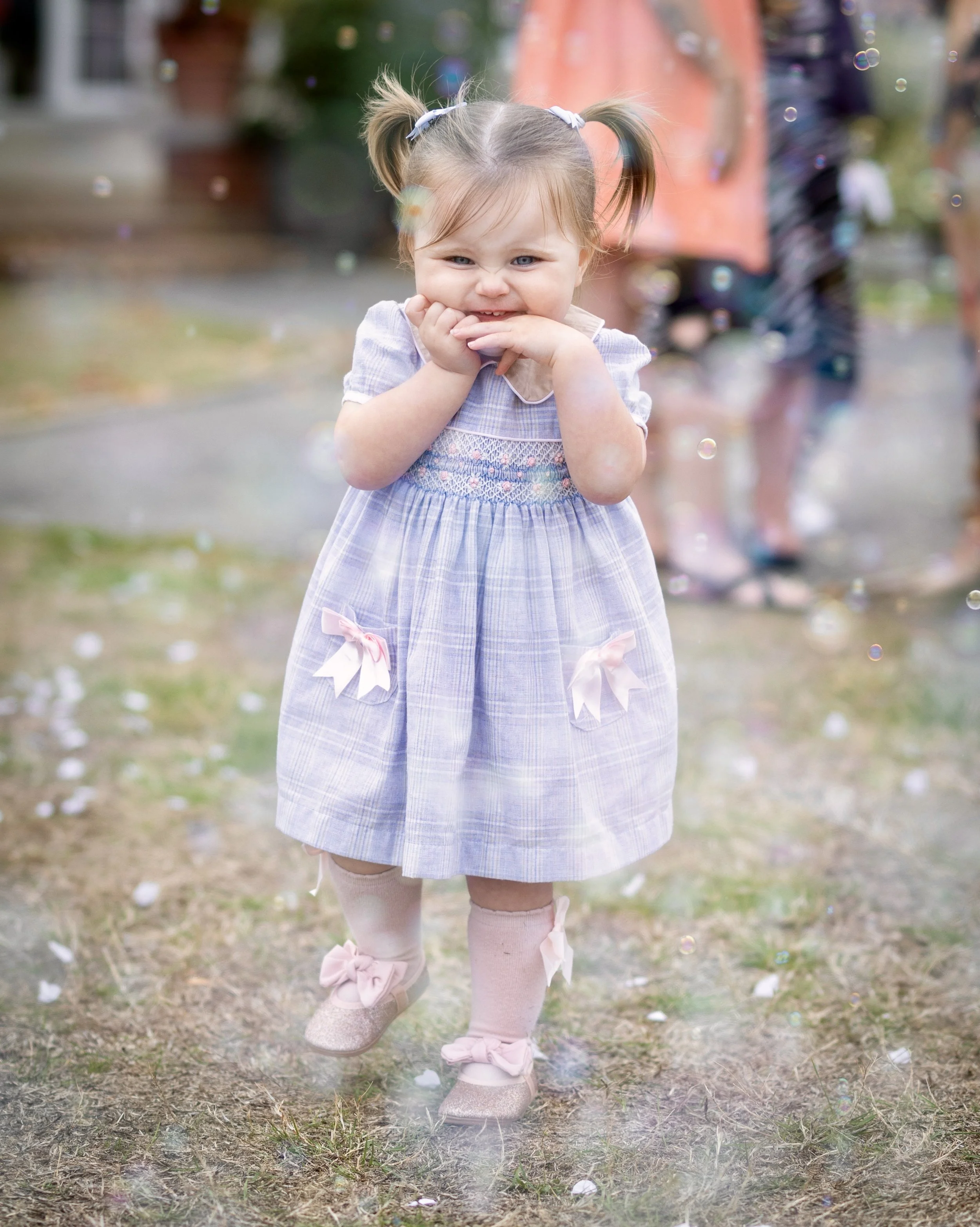 A young girl with light brown hair styled in pigtails, wearing a purple dress and pink tights with bows, is smiling and holding her hand to her mouth outdoors, with floating bubbles around her.