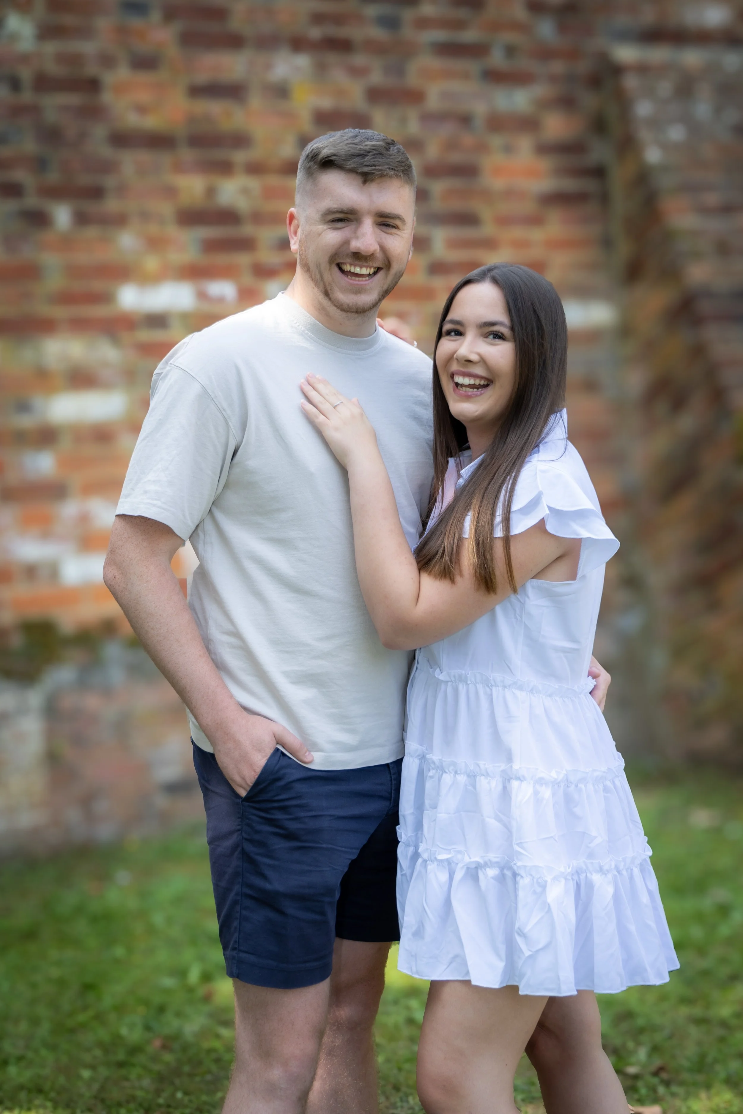 A smiling man in a light gray t-shirt and shorts standing next to a woman in a white dress with ruffled sleeves and a tiered skirt, both standing outdoors in front of a brick wall.