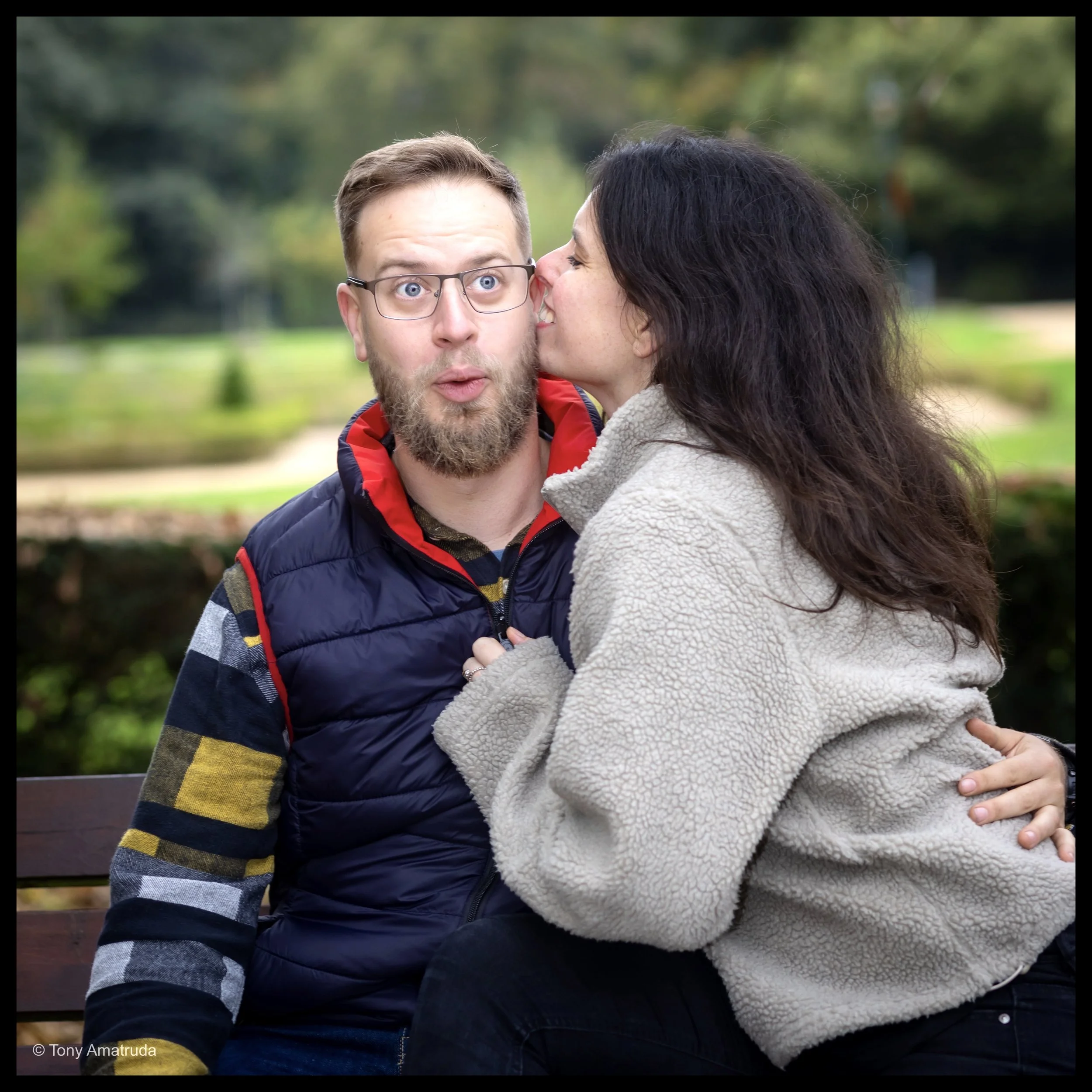 A woman with long dark hair is kissing a man with glasses, a beard, and light brown hair on the cheek while outdoors in a park with green trees in the background.