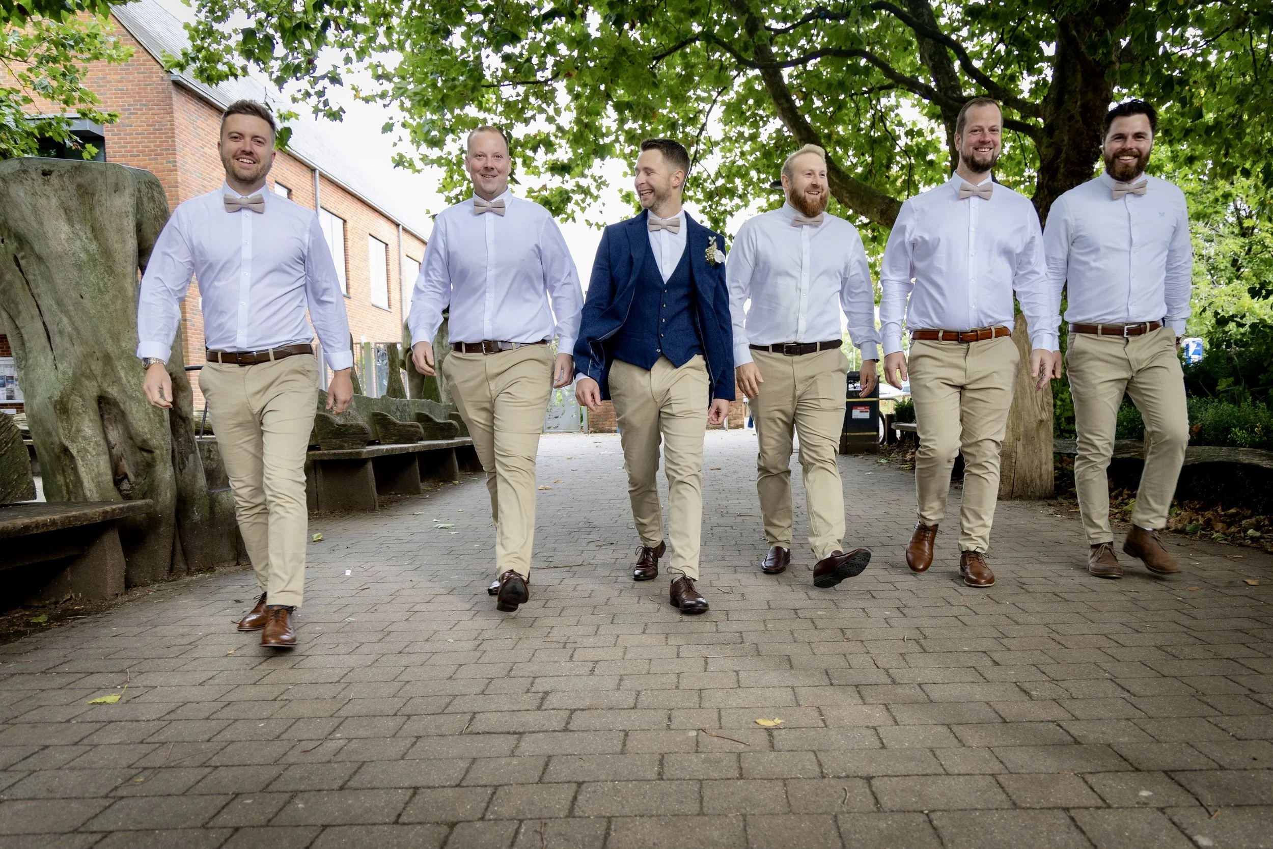 Six men walking outdoors under a large leafy tree, dressed in formal attire with beige pants, white shirts, and bow ties. One man in the center is wearing a blue suit jacket and has a boutonniere on his lapel. They are smiling and appear to be celebr